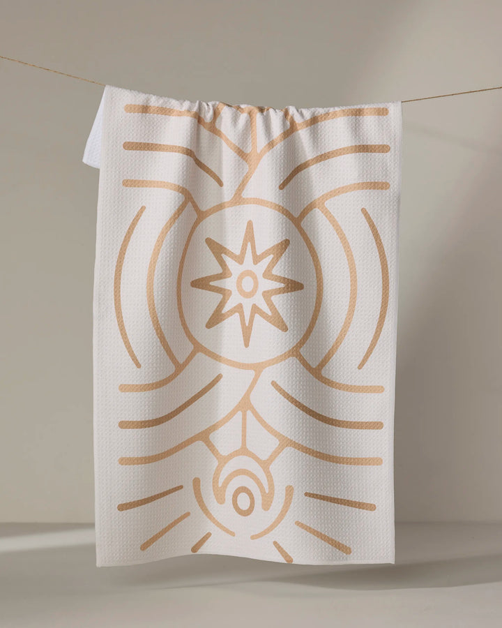 The Lunar tea towel by Adam Nienow features a recycled design with beige abstract stars and lines, hanging stylishly against a neutral background to bring sustainability and elegance to your kitchen.