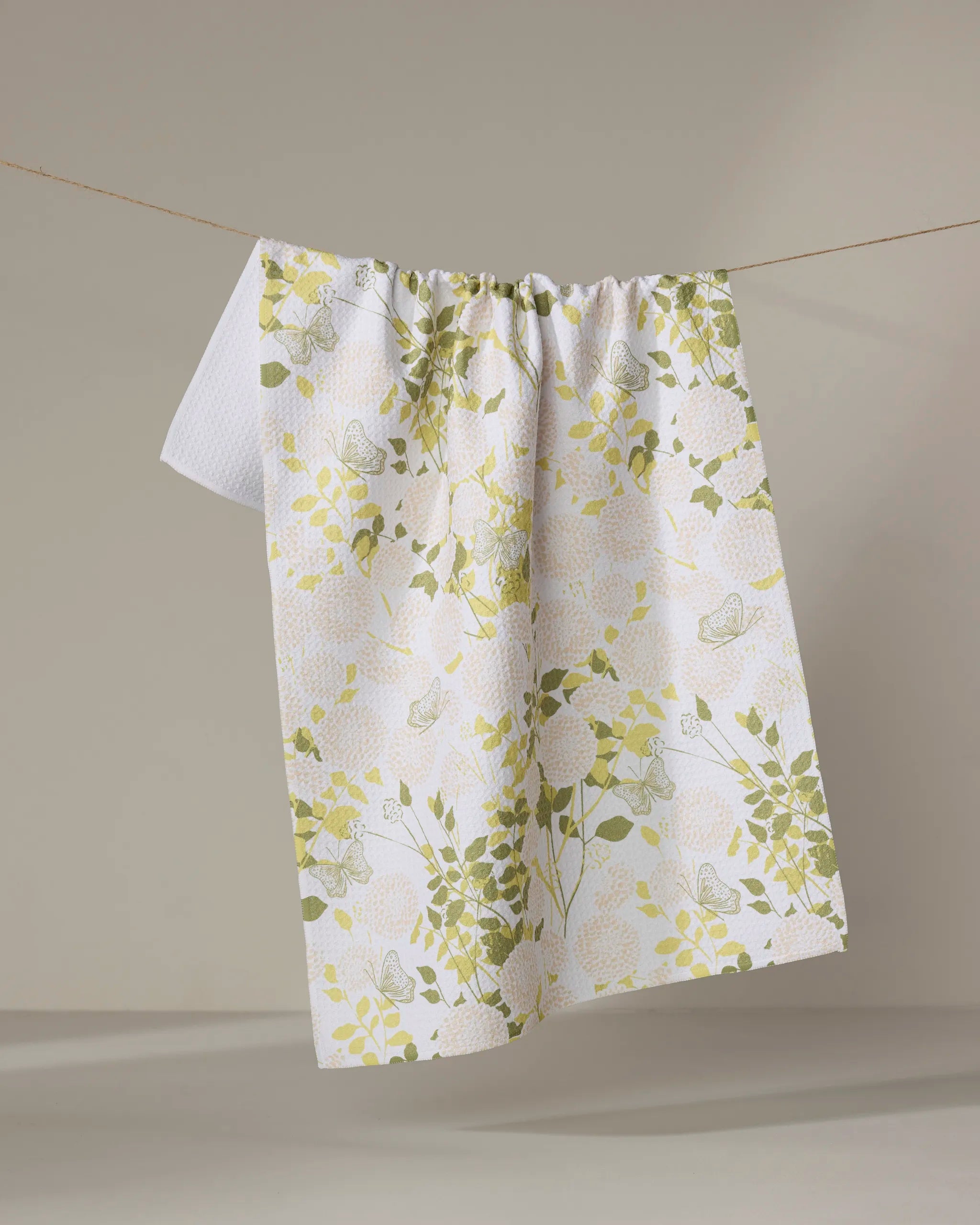 The Lush Garden tea towel by Second Nature Studio, featuring yellow and green floral and butterfly designs on white recycled fabric, hangs on a clothesline against a neutral background.