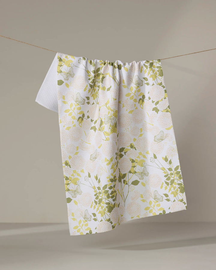The Lush Garden tea towel by Second Nature Studio, featuring yellow and green floral and butterfly designs on white recycled fabric, hangs on a clothesline against a neutral background.