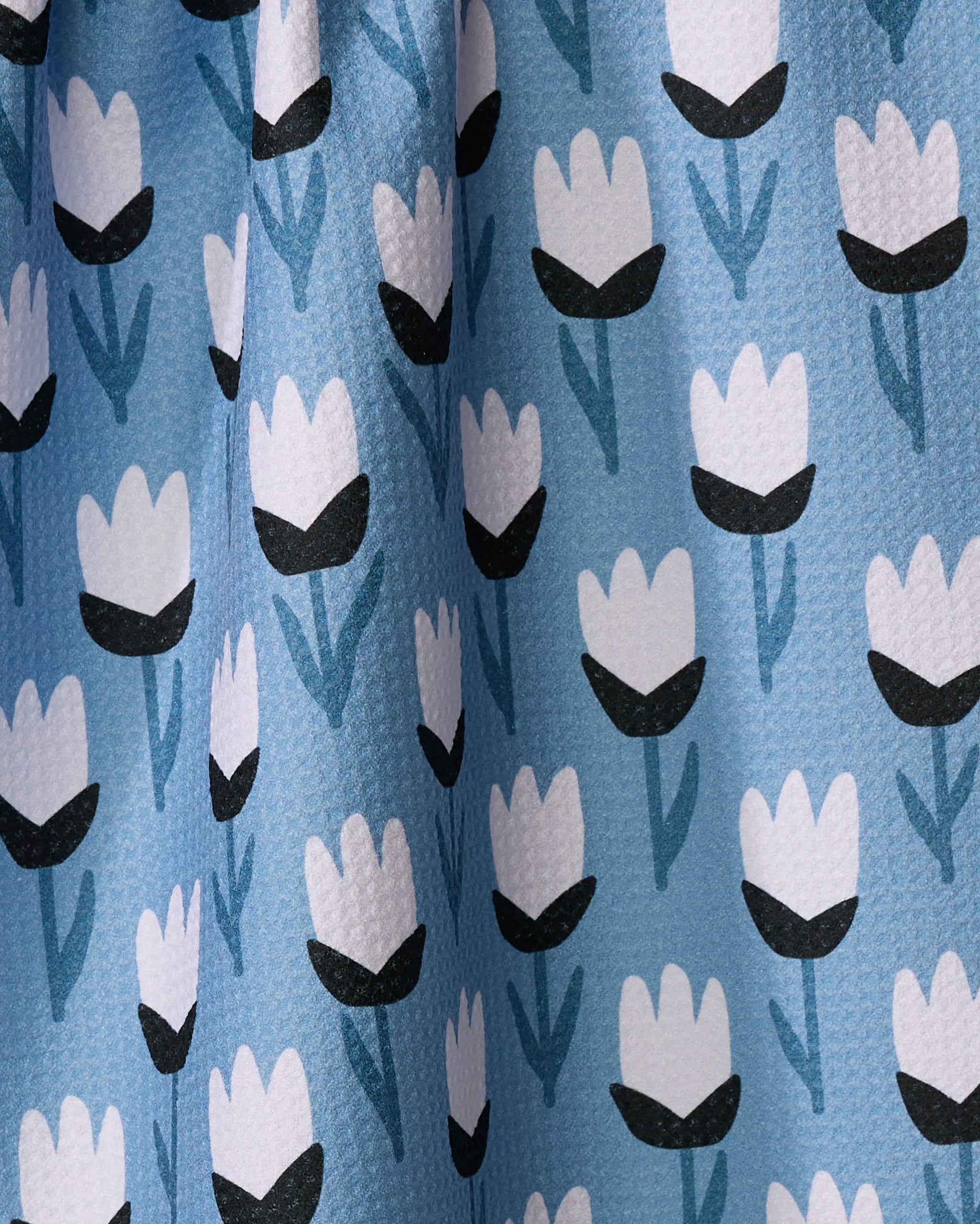 A close-up of the Lush Meadow kitchen towel by Anette Heiberg shows textured light blue fabric with a charming pattern of white tulip flowers, black centers, and blue stems—an elegant touch for any kitchen.