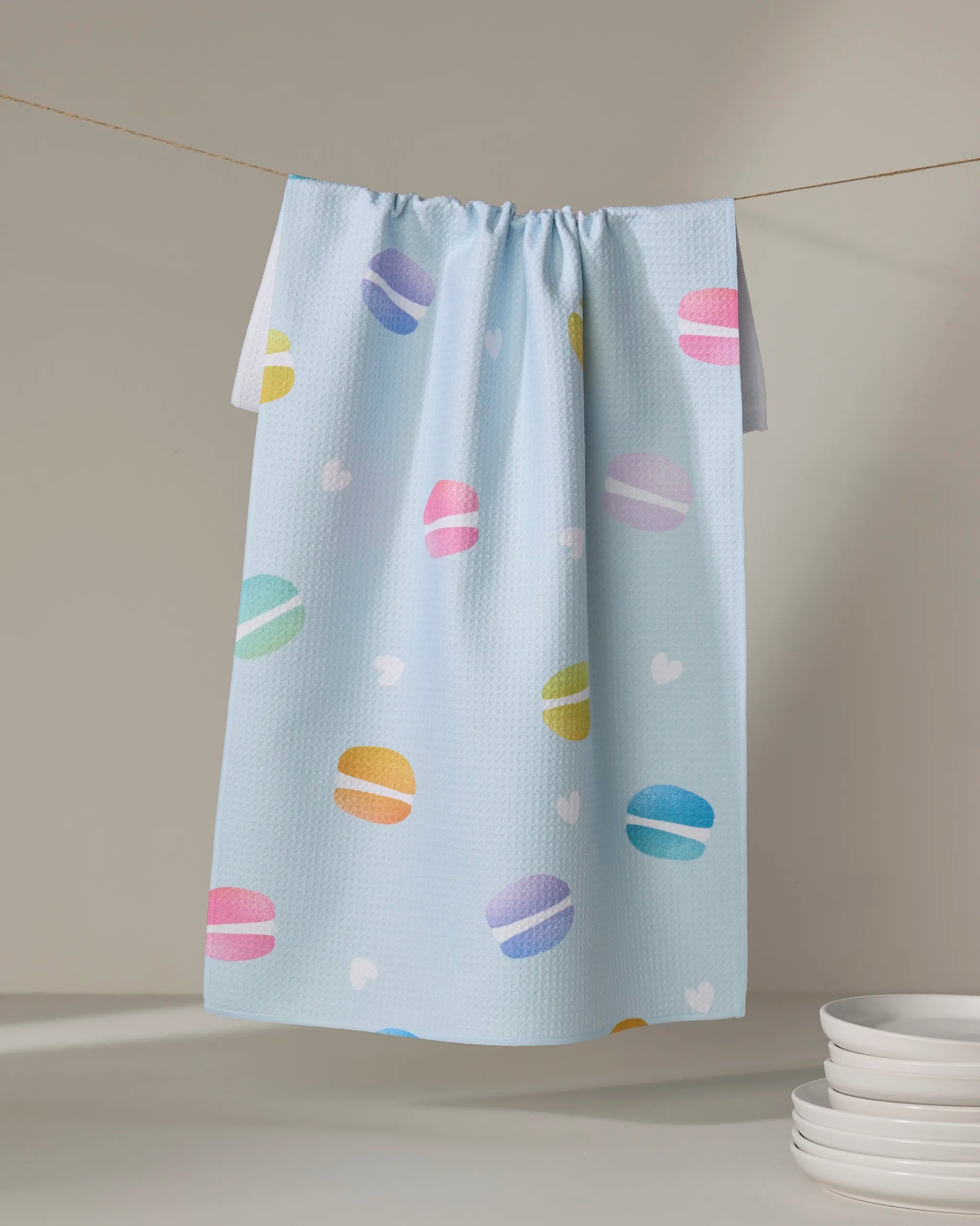 The Mac Flip tea towel by Nora's Macarons, in light blue with colorful macarons and white hearts, hangs on a clothesline next to stacked white plates in the bottom right corner.