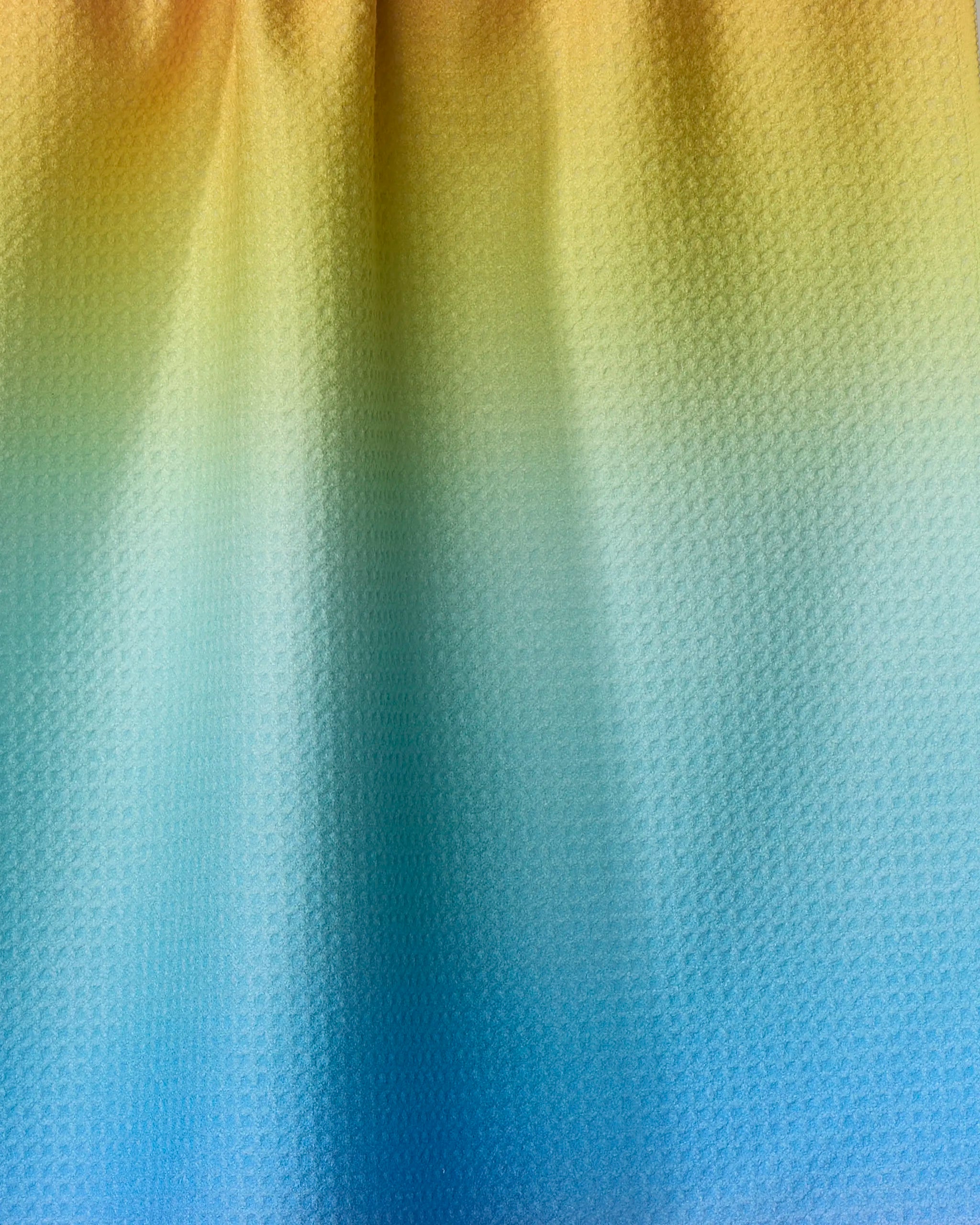 The Mac Joy tea towel by Nora's Macarons is crafted from recycled materials and features a textured design with a soft ombre gradient that shifts from yellow at the top to blue at the bottom.