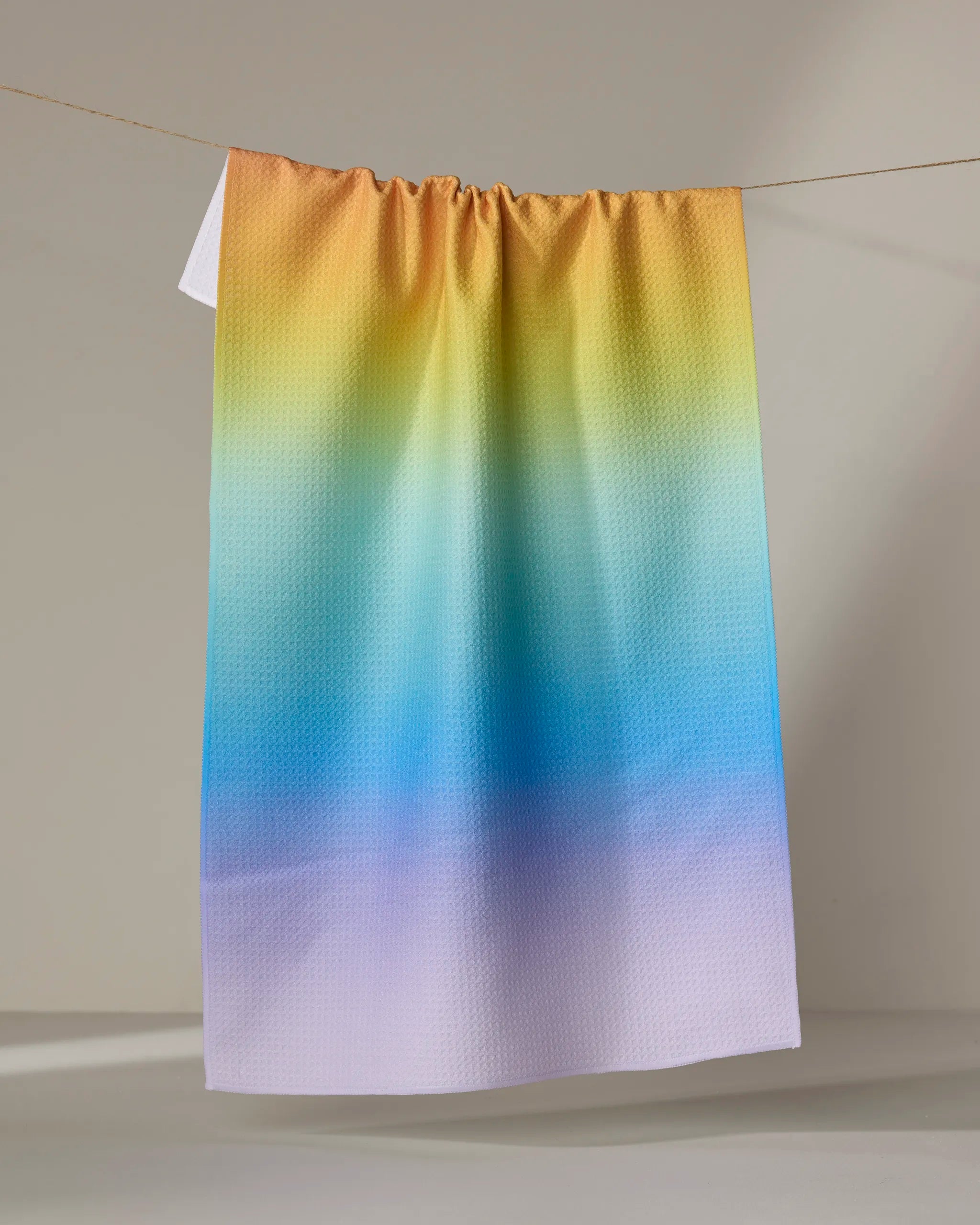 The Mac Joy tea towel by Nora's Macarons, featuring a yellow, green, blue, and purple gradient pattern on recycled fabric, hangs from a string against a neutral background.