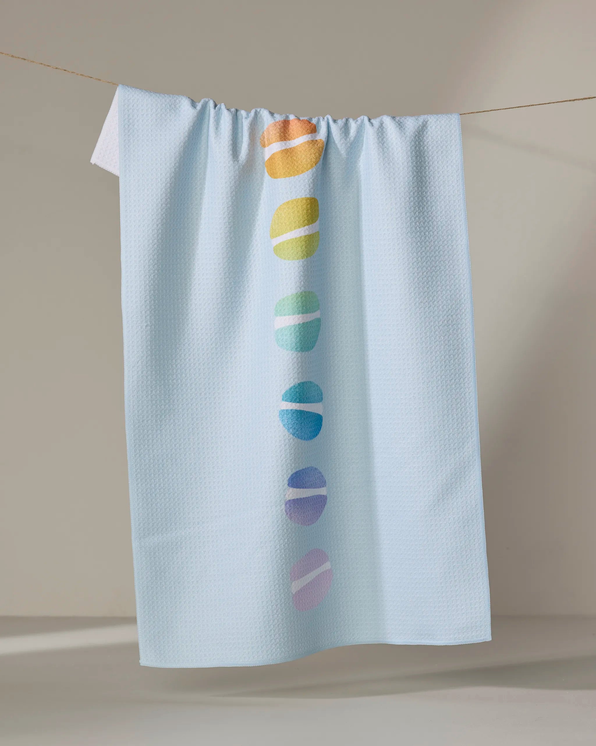 A light blue textured kitchen towel from Nora's Macarons, called "Mac Love," hangs on a line and features seven colorful oval shapes in orange, yellow, green, blue, and purple down the center.