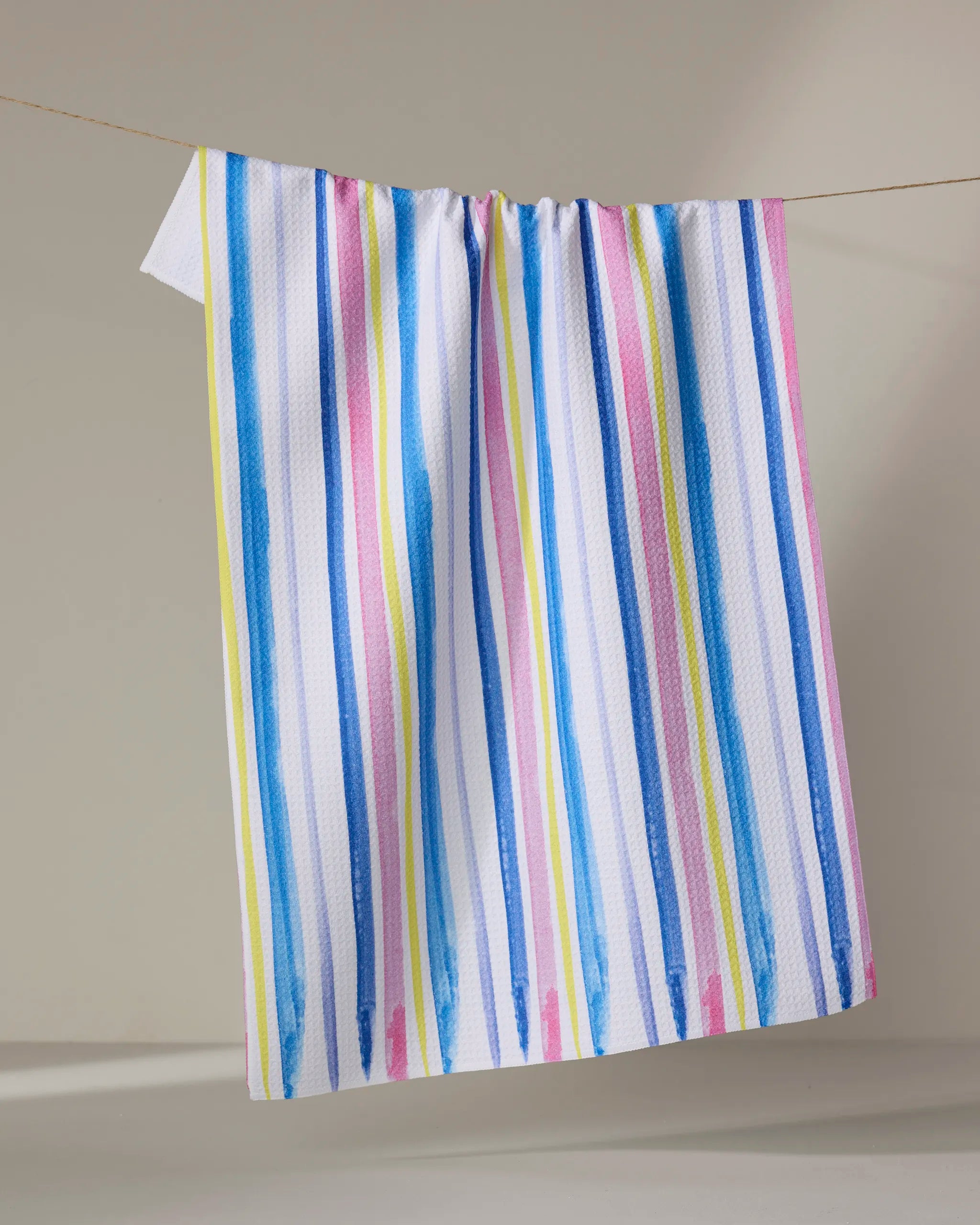 The Macarons in Paris Stripe tea towel by Travel Write Draw features eco-friendly, colorful vertical stripes and hangs on a clothesline against a neutral background.