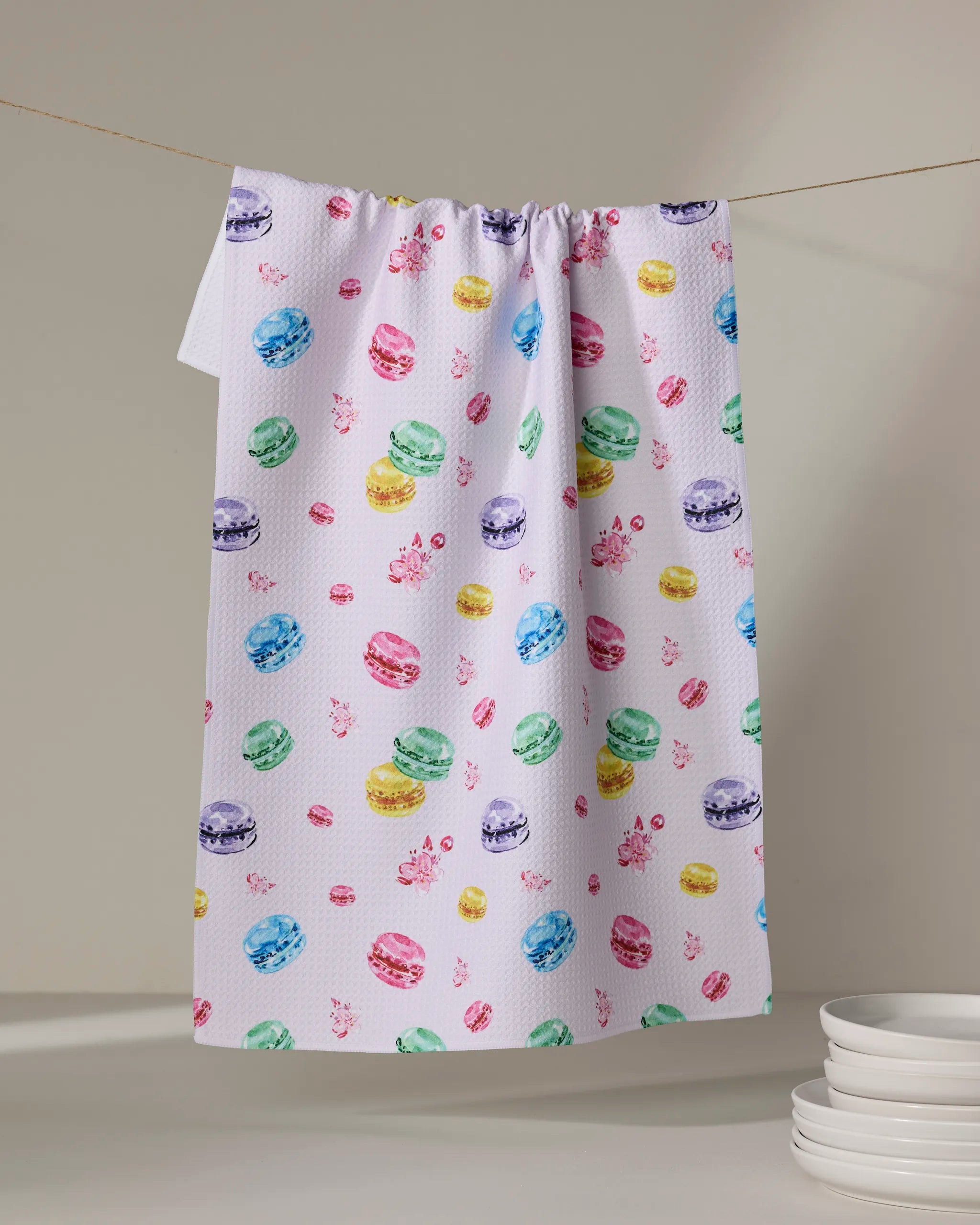 The "Macarons in Paris" tea towel by Travel Write Draw by Meagan Morrison, made from recycled materials, features a vibrant macaron pattern and hangs against a neutral background with white plates nearby for an eco-friendly touch.