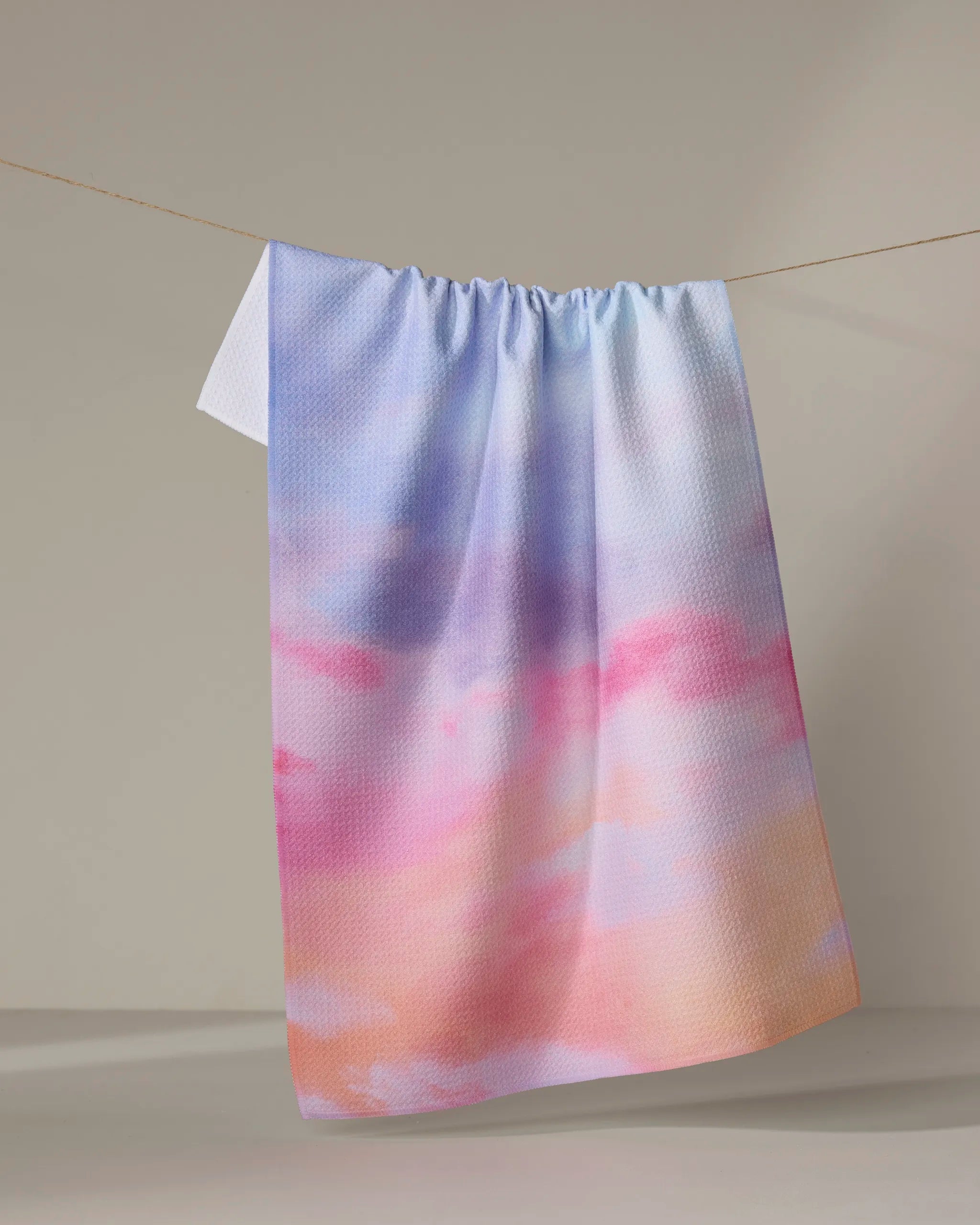 The "Magic In The Sky" dish towel by Stefanie Bales, featuring absorbent fabric in a pastel gradient of blue, pink, and peach hues, hangs on a clothesline against a neutral background.