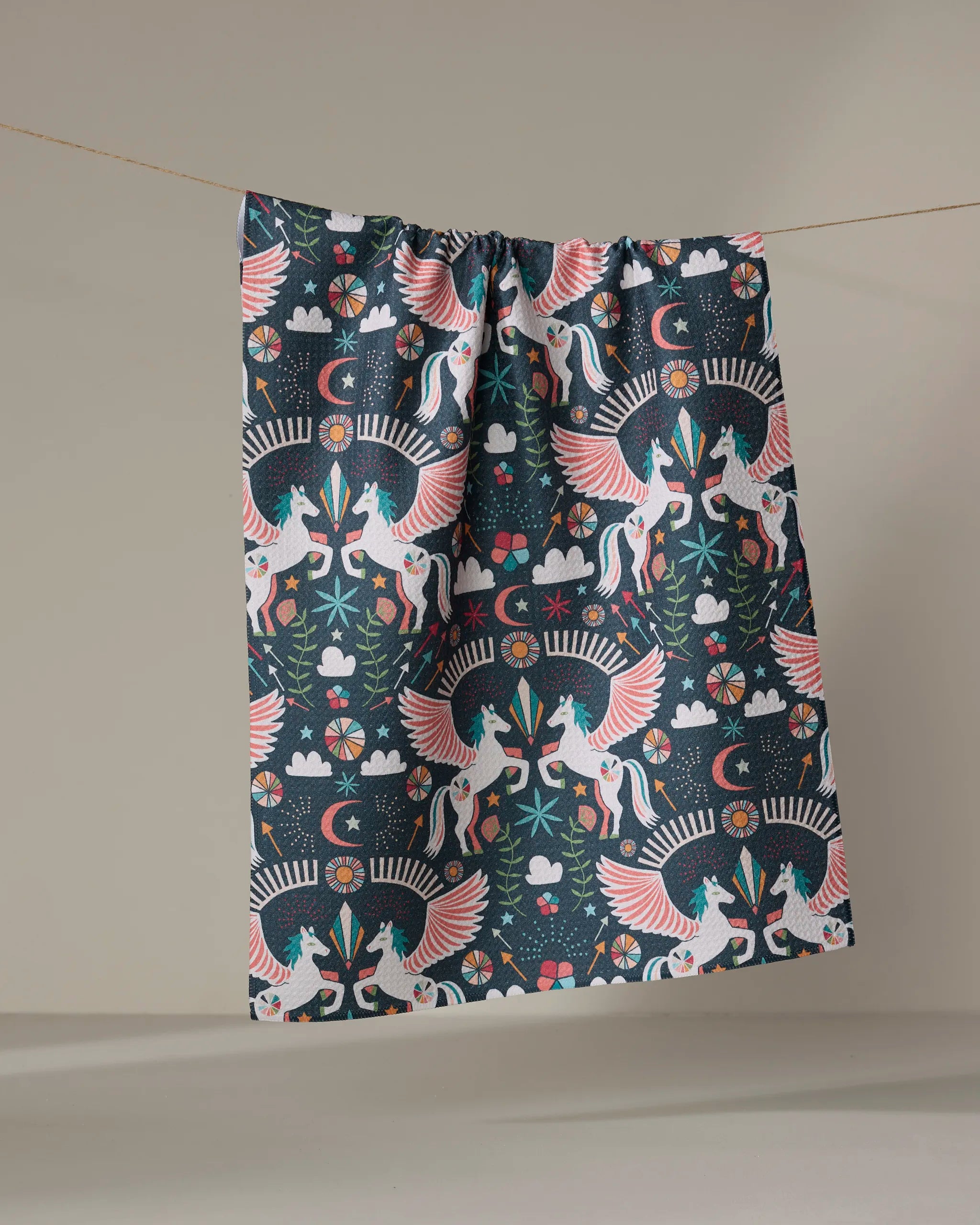 The Magical Moments tea towel by Jessica Swift features white unicorns with pink wings among flowers, stars, clouds, and plants on a dark background—a whimsical, quick-drying fabric panel perfect for magical kitchens.