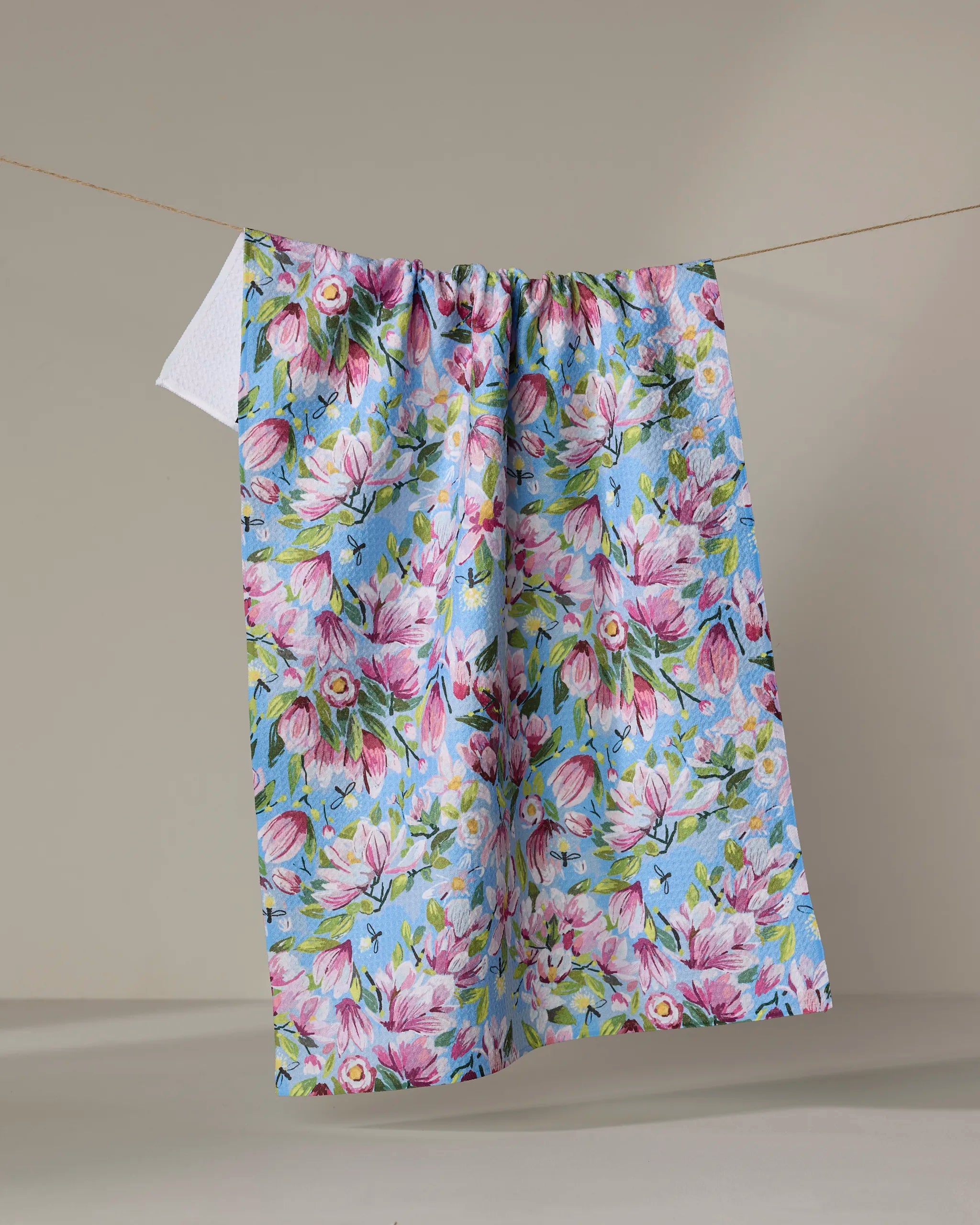 The "Magnolia" tea towel by Elisabeth Wing, featuring a pink and white floral design on blue and made from recycled materials, hangs on a clothesline against a neutral background.