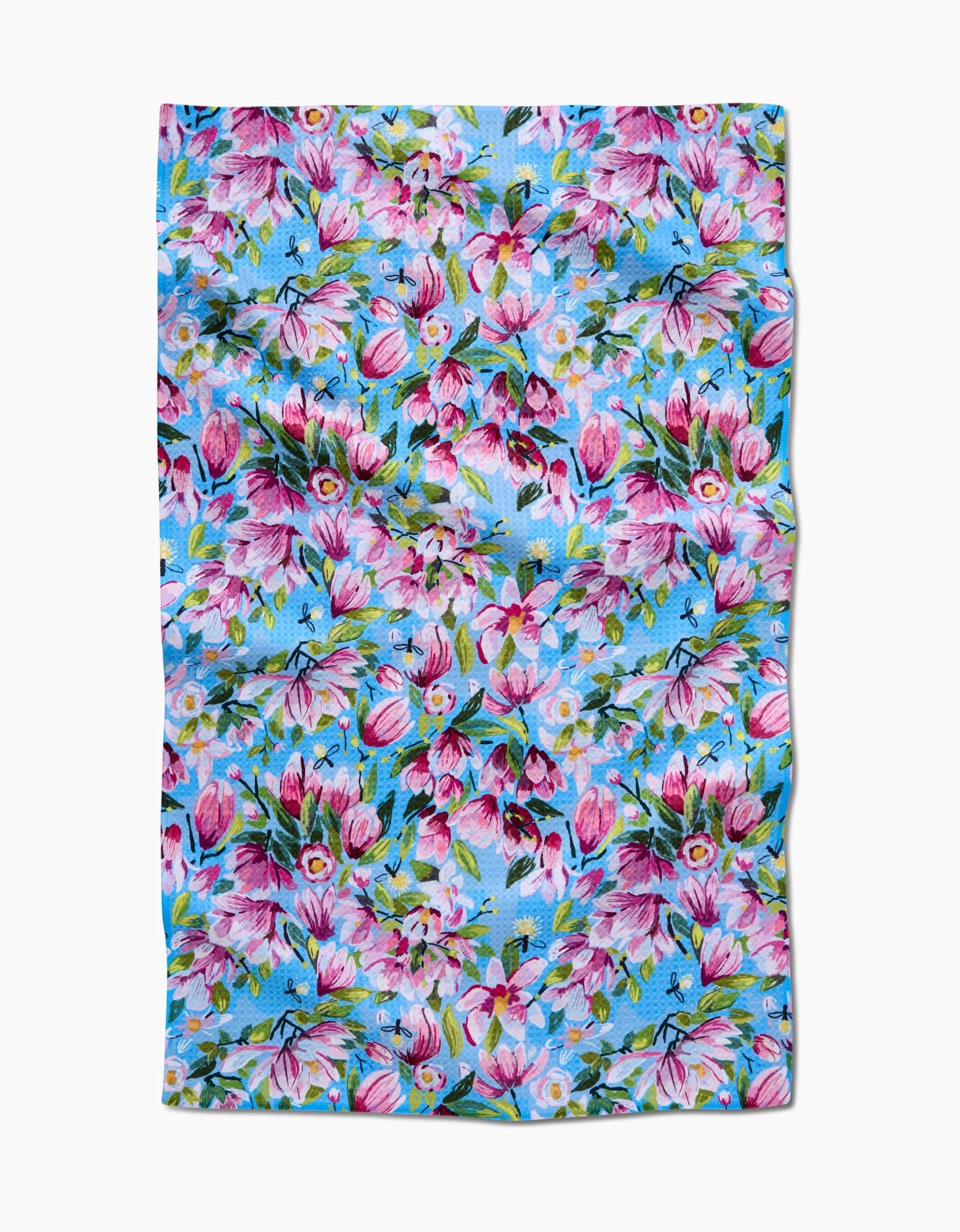 The Magnolia Tea Towel by Elisabeth Wing features pink and white flowers with green leaves on a bright blue background. Made from ultra-absorbent Geoweave fabric, it is rectangular and shown slightly wrinkled, laid flat.