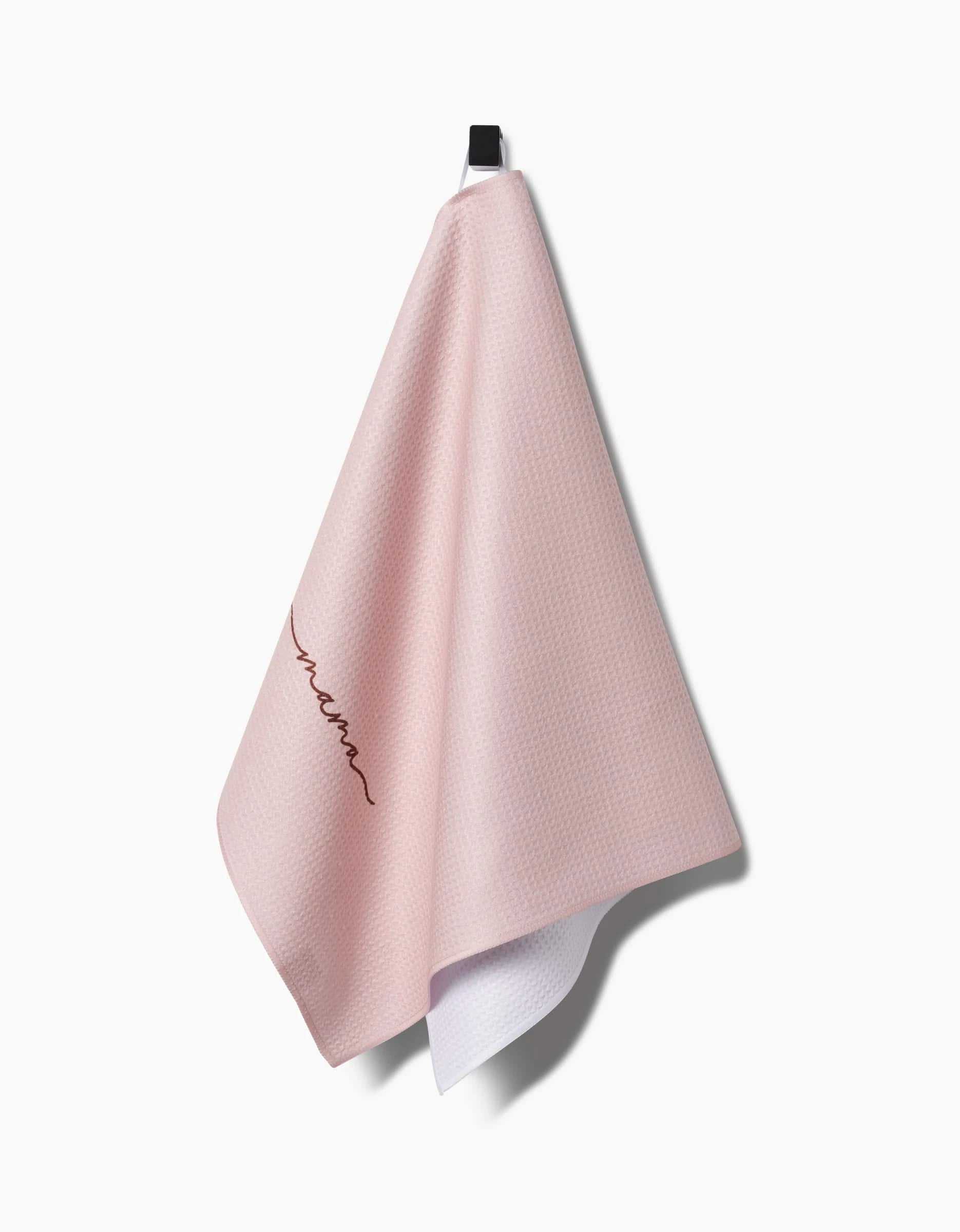 The Mama towel by Kailee Wright is a light pink, ultra-absorbent Geoweave towel with a subtle waffle texture and embroidered cursive script on one side, shown hanging from a black hook against a white background.