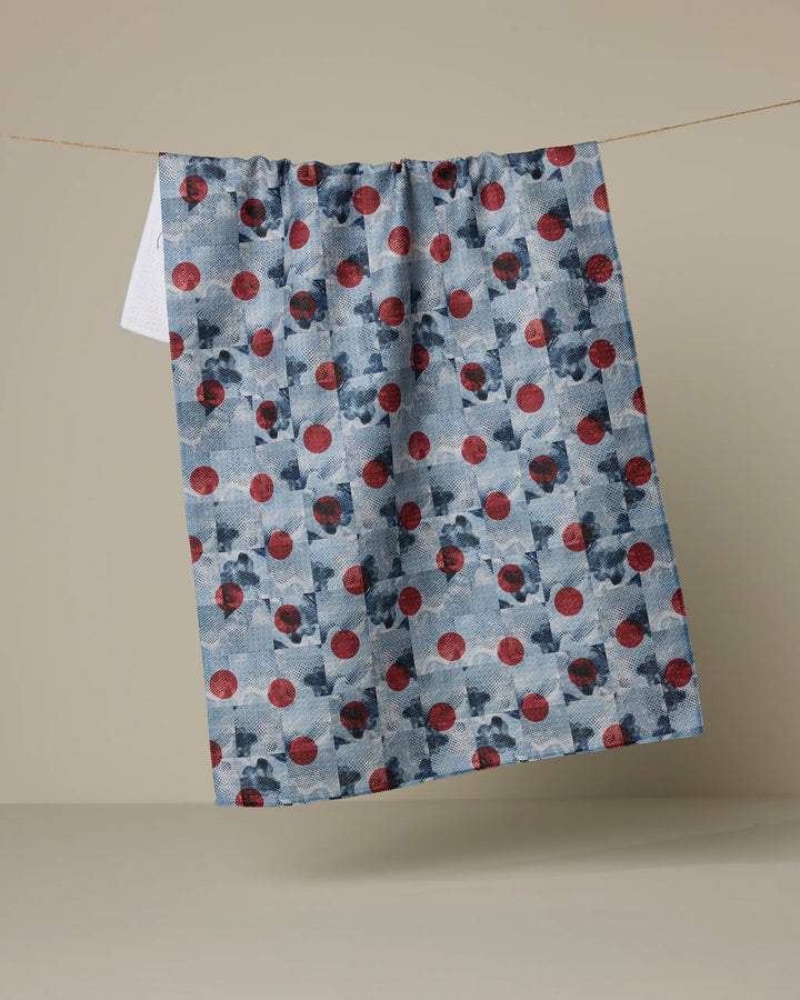 The Manga Dot kitchen tea towel by Geometry features dark blue shapes and red circles on blue fabric, hanging from a string against a plain beige background.