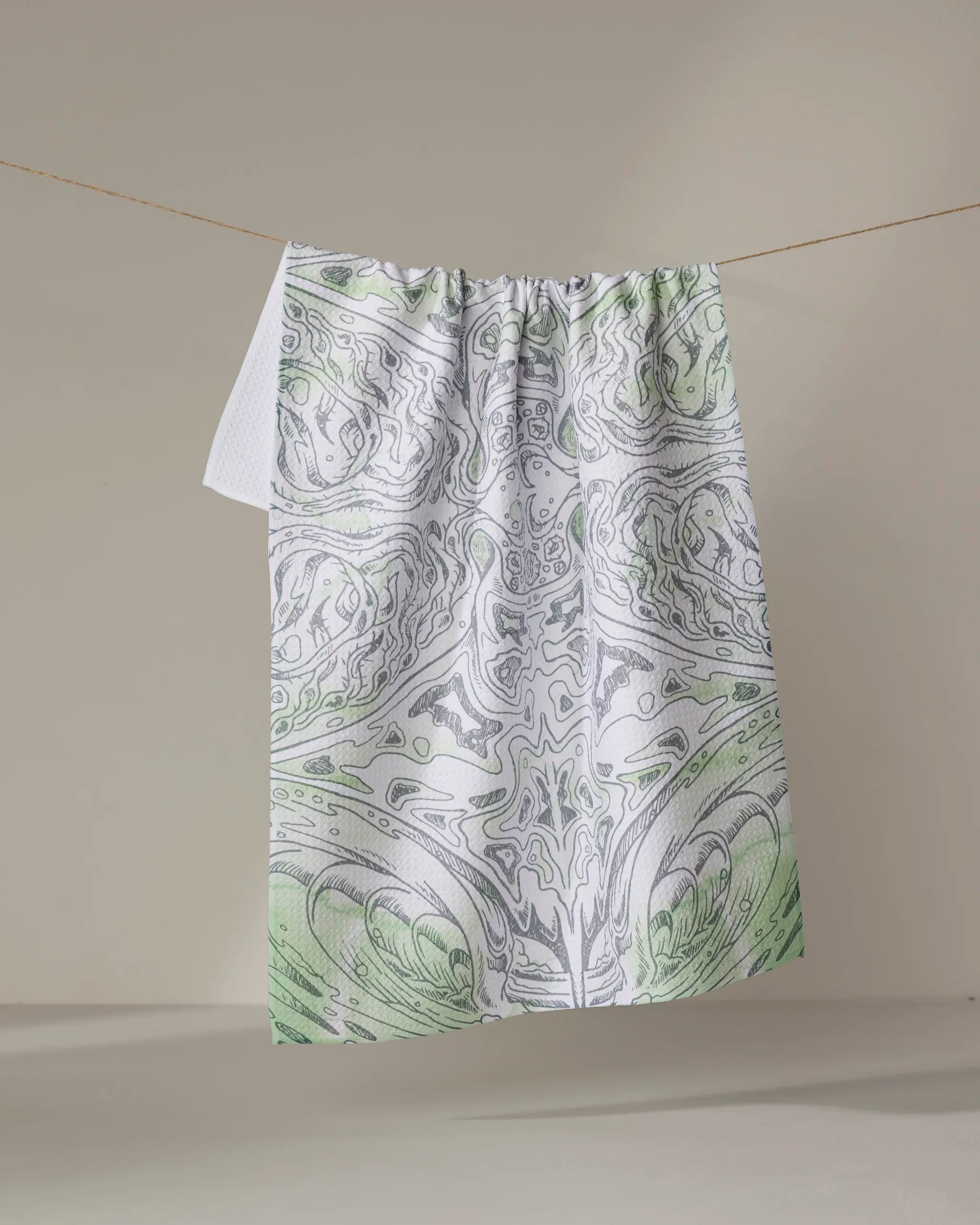 The Skye Walker "Manta" tea towel, made from recycled materials, displays an intricate green and gray abstract pattern and hangs against a neutral background.