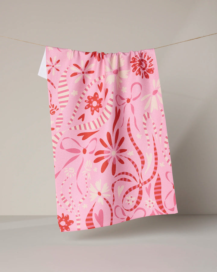 The Geometry Marcela tea towel, made from recycled materials, displays playful red, pink, and white floral and ribbon patterns and hangs on a clothesline against a light gray background.