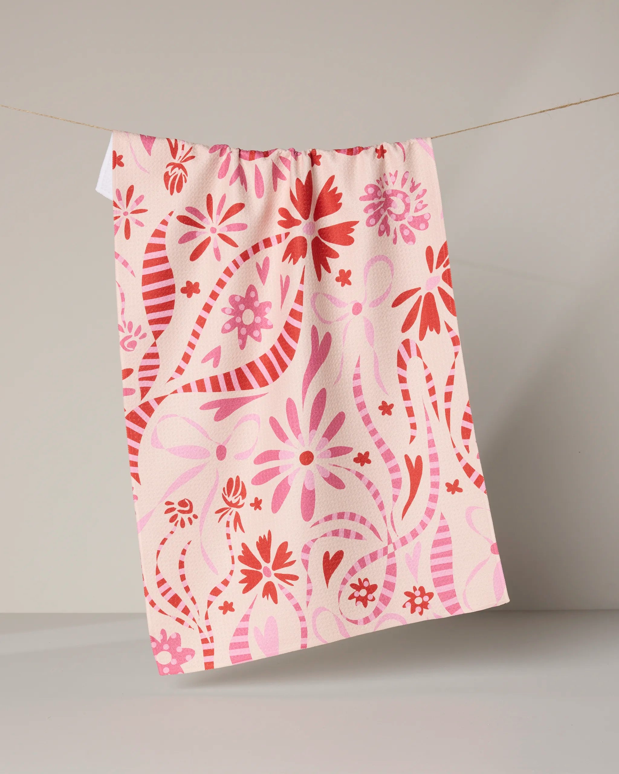 The Geometry Maricela tea towel, featuring quick-drying fabric with pink and red floral and striped designs, hangs on a string against a neutral background.