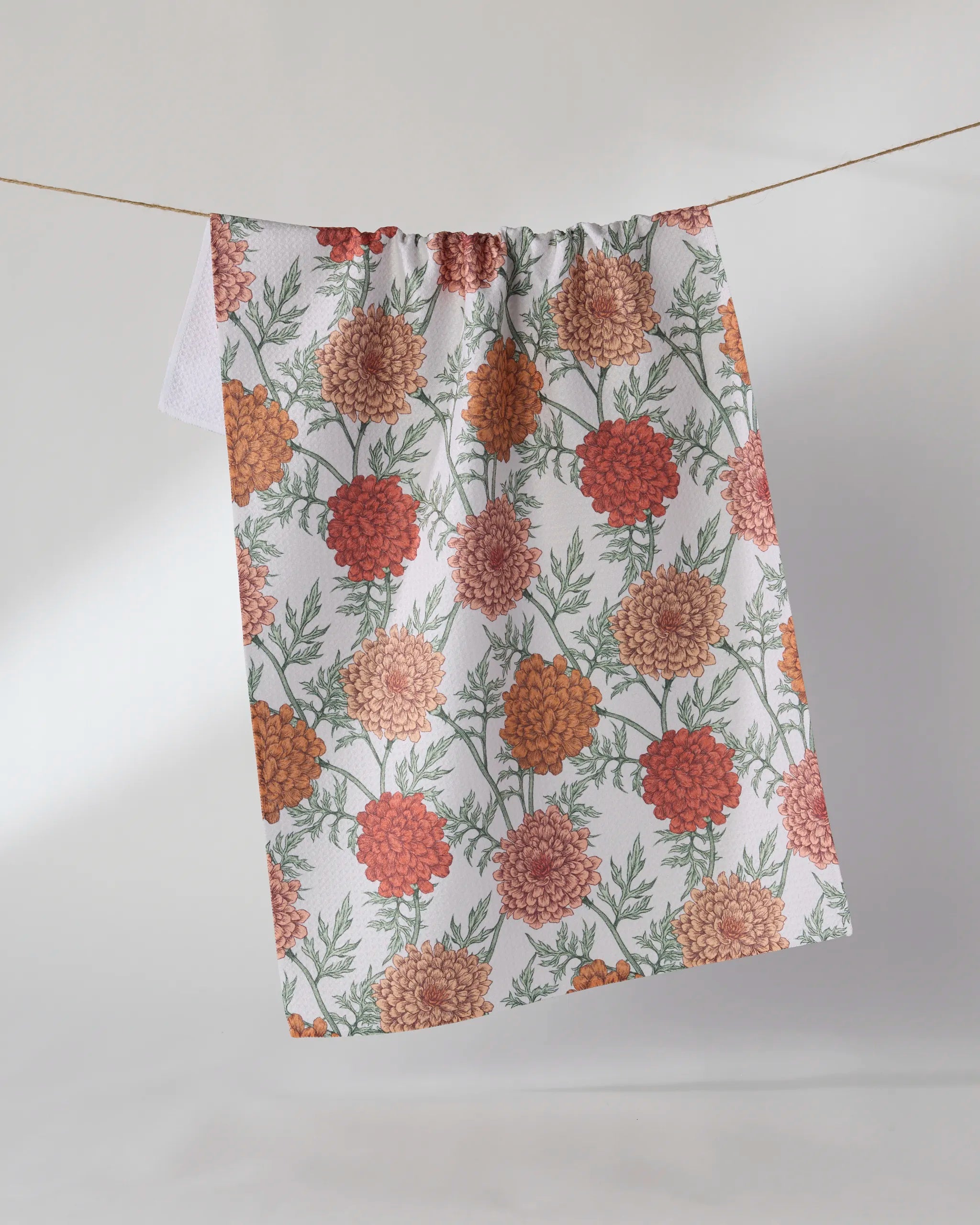 The Geometry Marigold Manor tea towel, made from recycled materials, features orange and peach marigolds with green leaves on white fabric and hangs on a clothesline against a plain light background.
