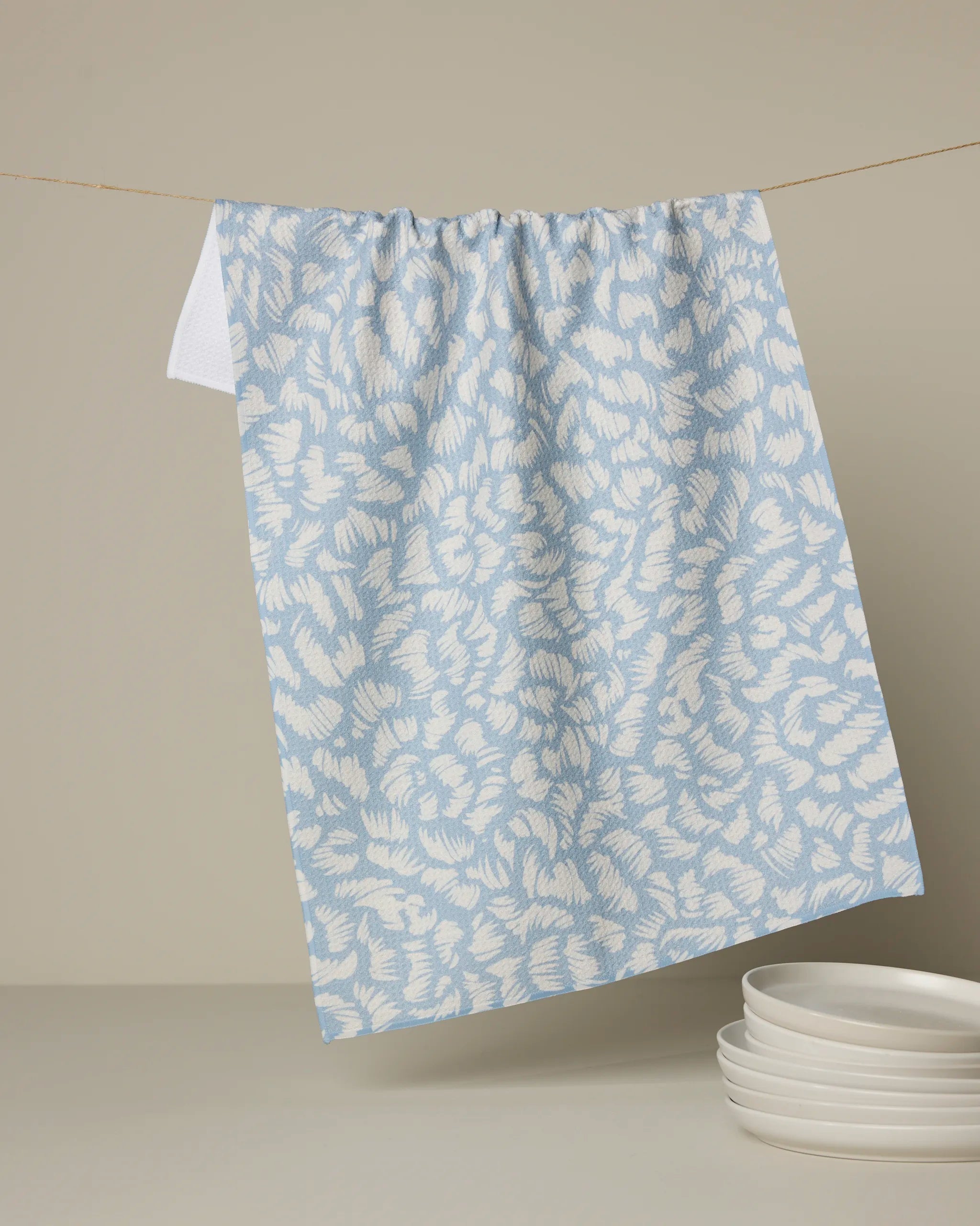 The Geometry Marine Haze tea towel, featuring a light blue and white pattern and made from recycled materials, hangs on a string against a beige background with a stack of white plates below.