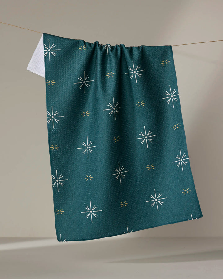 A Twig + Spark "Marine Navigation" tea towel, made from dark green Geoweave fabric with white starbursts and yellow accents, hangs on a line against a neutral background.