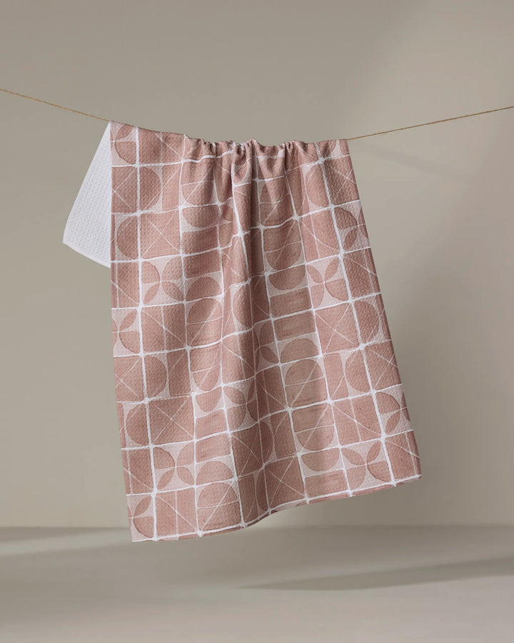 The Mariposa towel by Studio Jeannot, featuring a quick-drying beige fabric with a white geometric pattern, hangs from a string against a plain, neutral background.