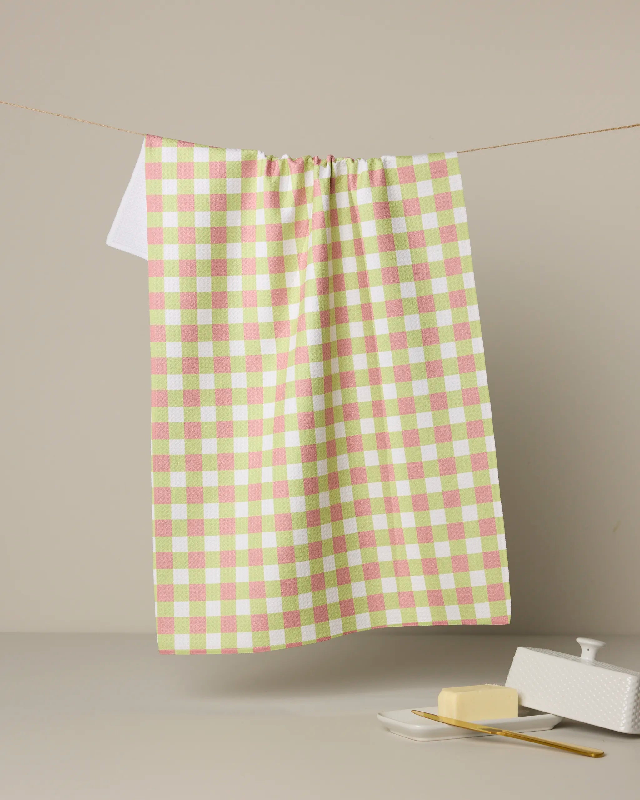 The Geometry Mariquita Gingham kitchen towel in pastel pink, green, and white hangs on a string against a beige background, with a butter dish, butter, and knife displayed underneath.