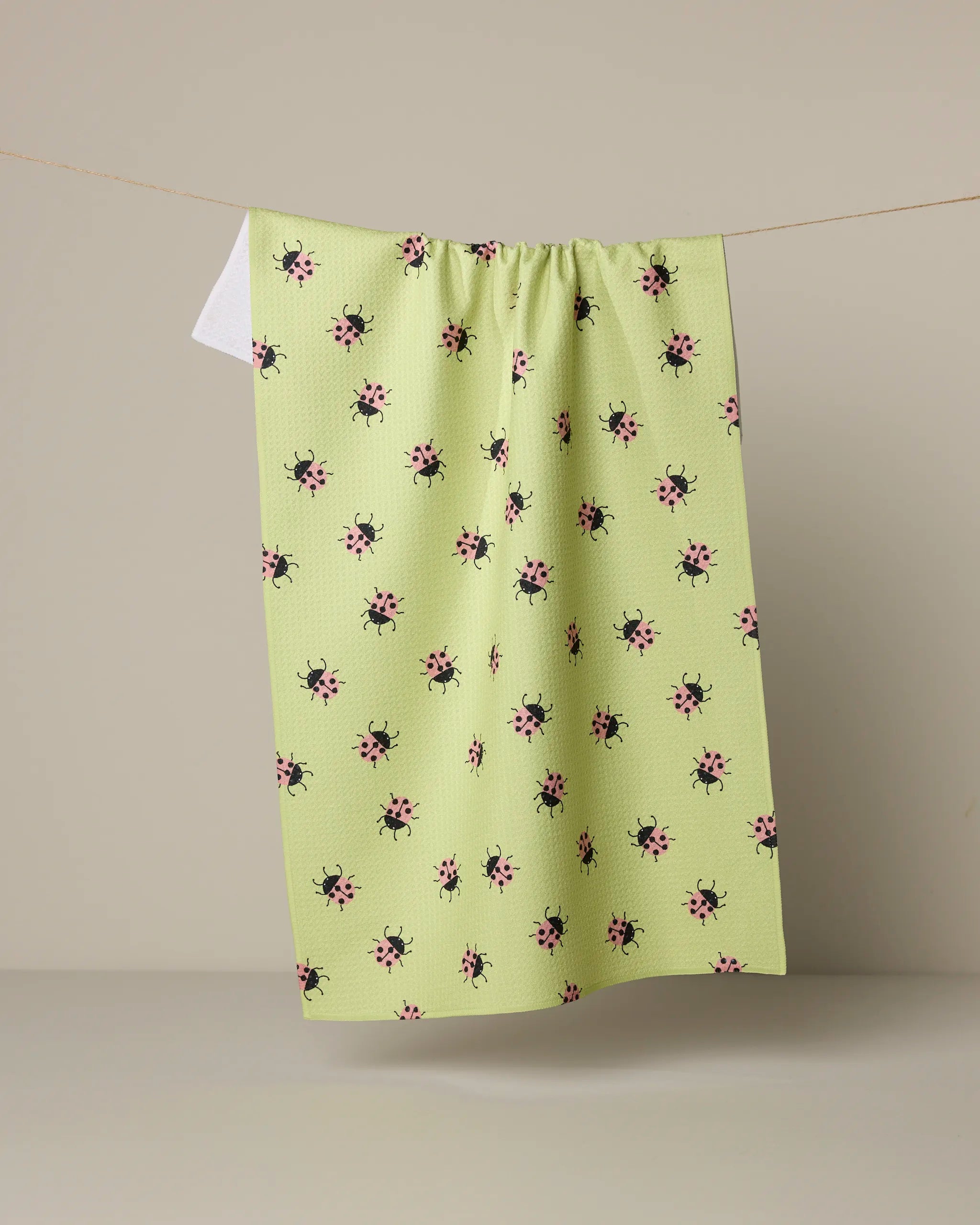 Geometry’s Mariquita tea towel, featuring a pattern of small pink and black ladybugs, hangs on a clothesline against a neutral background.