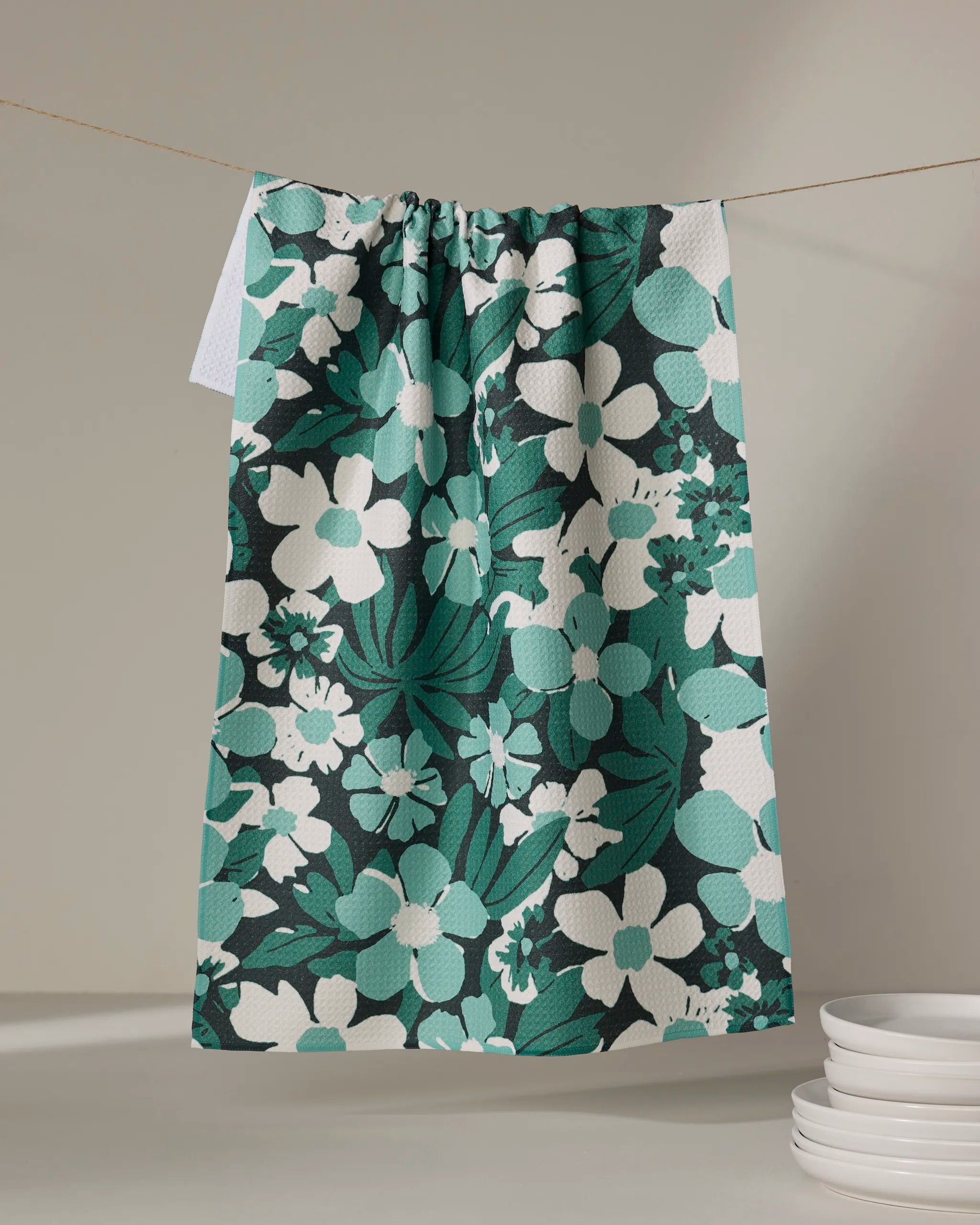 The Geometry Marlo Green tea towel, featuring a bold green and white floral pattern, dries quickly on a clothesline with a stack of white plates nearby.
