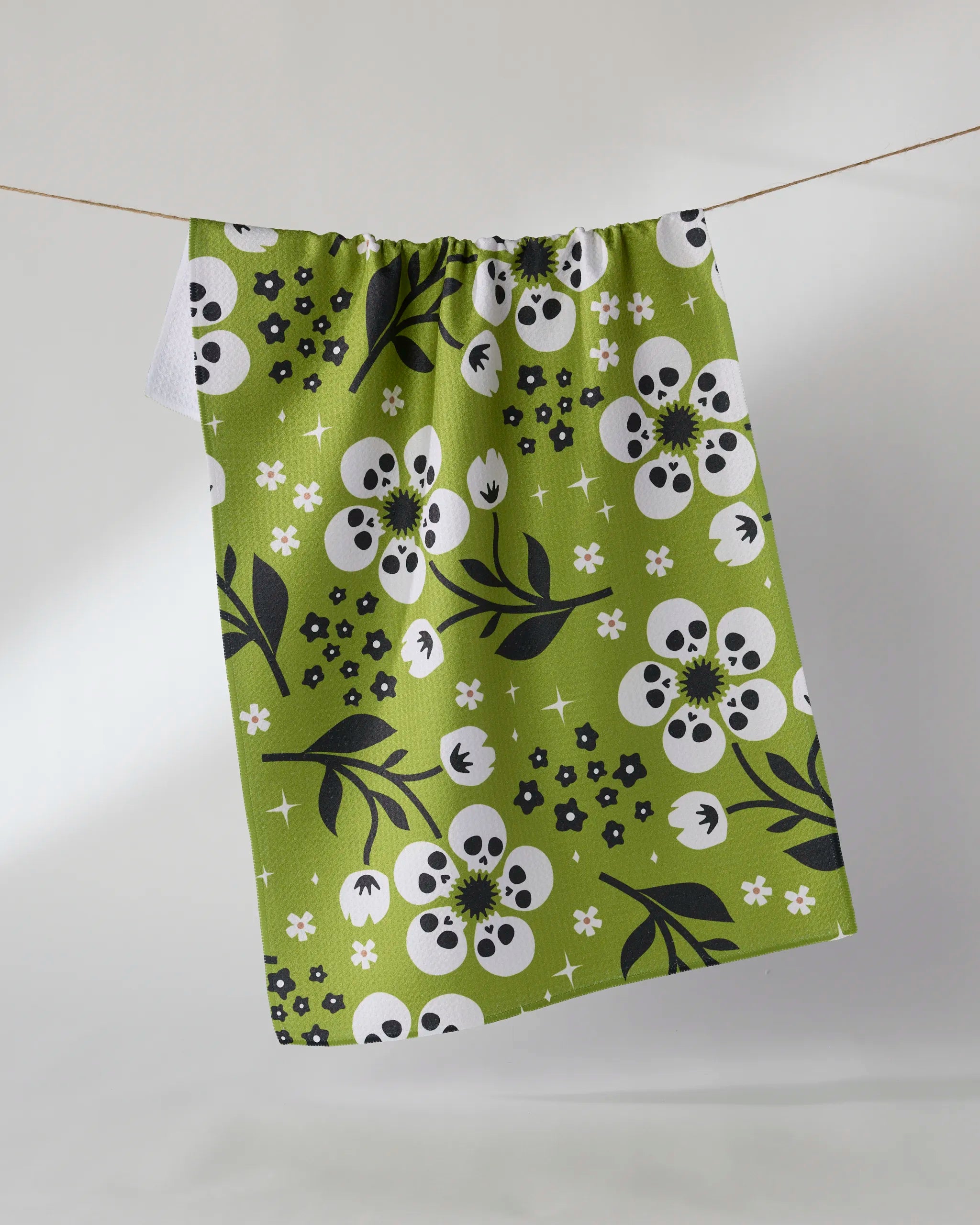 The Marrow Bloom tea towel by Geometry is made from recycled materials and features a playful pattern of white flowers with skull-like centers and black leaves on green, shown hanging against a plain light background.