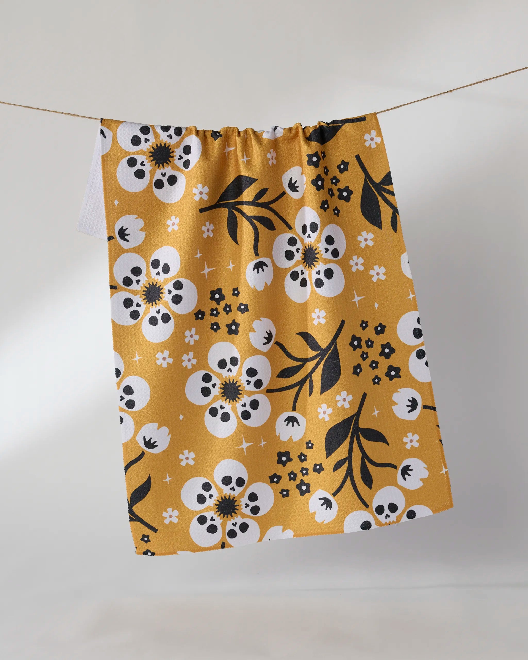 The Marrow Bloom tea towel by Geometry, made from recycled materials, showcases white flowers with black skull centers, black leaves, and floral accents on yellow fabric, hanging against a plain background.