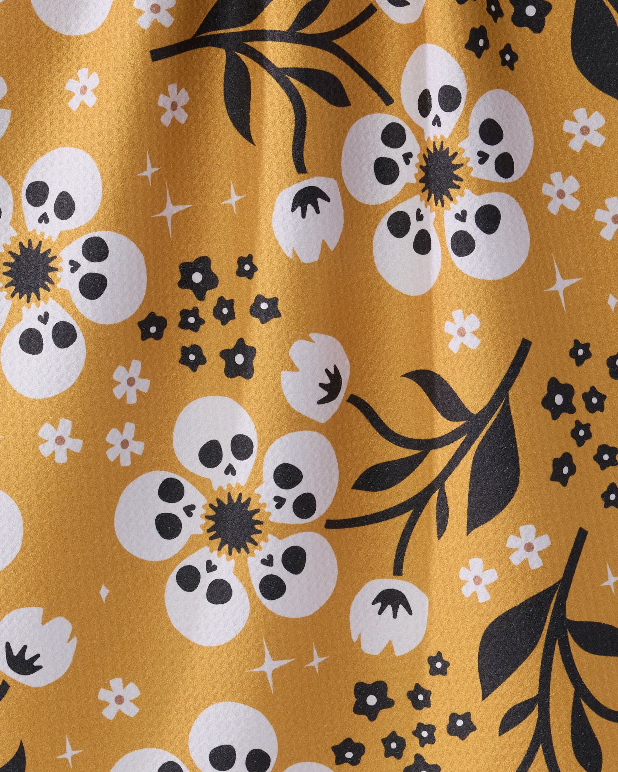 Geometry's Marrow Bloom tea towel features a whimsical yellow design with white skull-centered flowers, black leaves, and star accents. Made from recycled materials, its textured, wavy look adds charm to your kitchen.