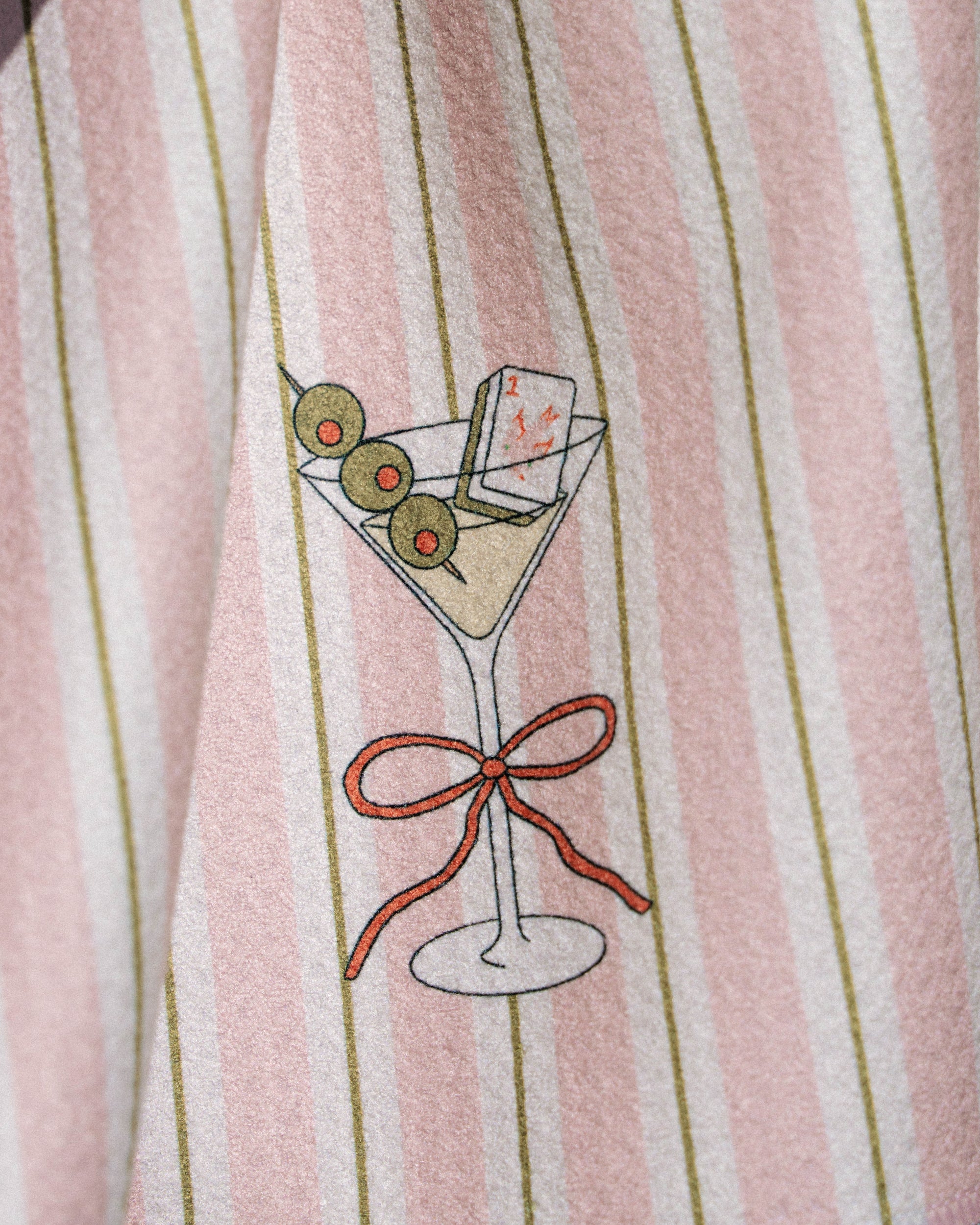 A closeup of Geometry’s Martini Stripe ultra-absorbent towel, showcasing pink and white stripes with a martini glass illustration featuring three olives, a playing card, and a red bow on the stem.