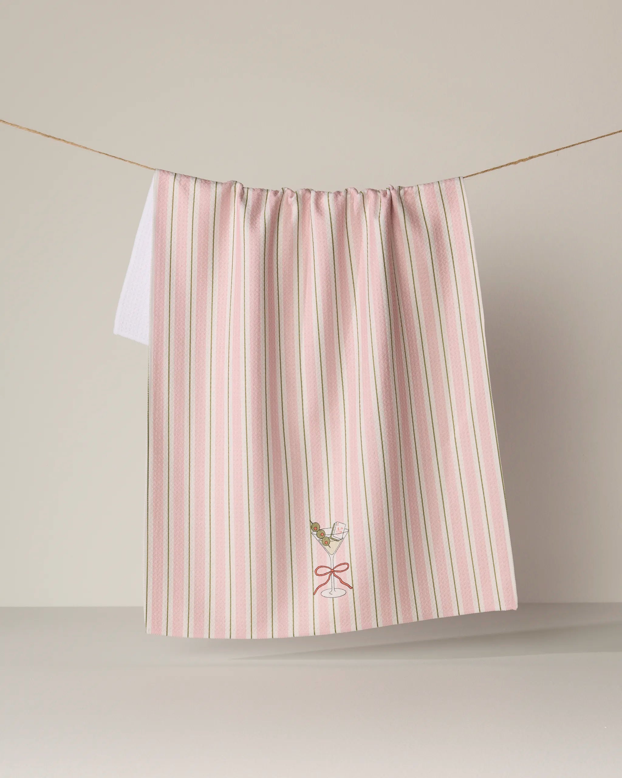 The Martini Stripe kitchen tea towel from Geometry features pink and white stripes, is ultra-absorbent, and made from recycled materials. A small embroidered martini glass with an olive adds a playful touch. The towel is displayed hanging on a string against a plain background.