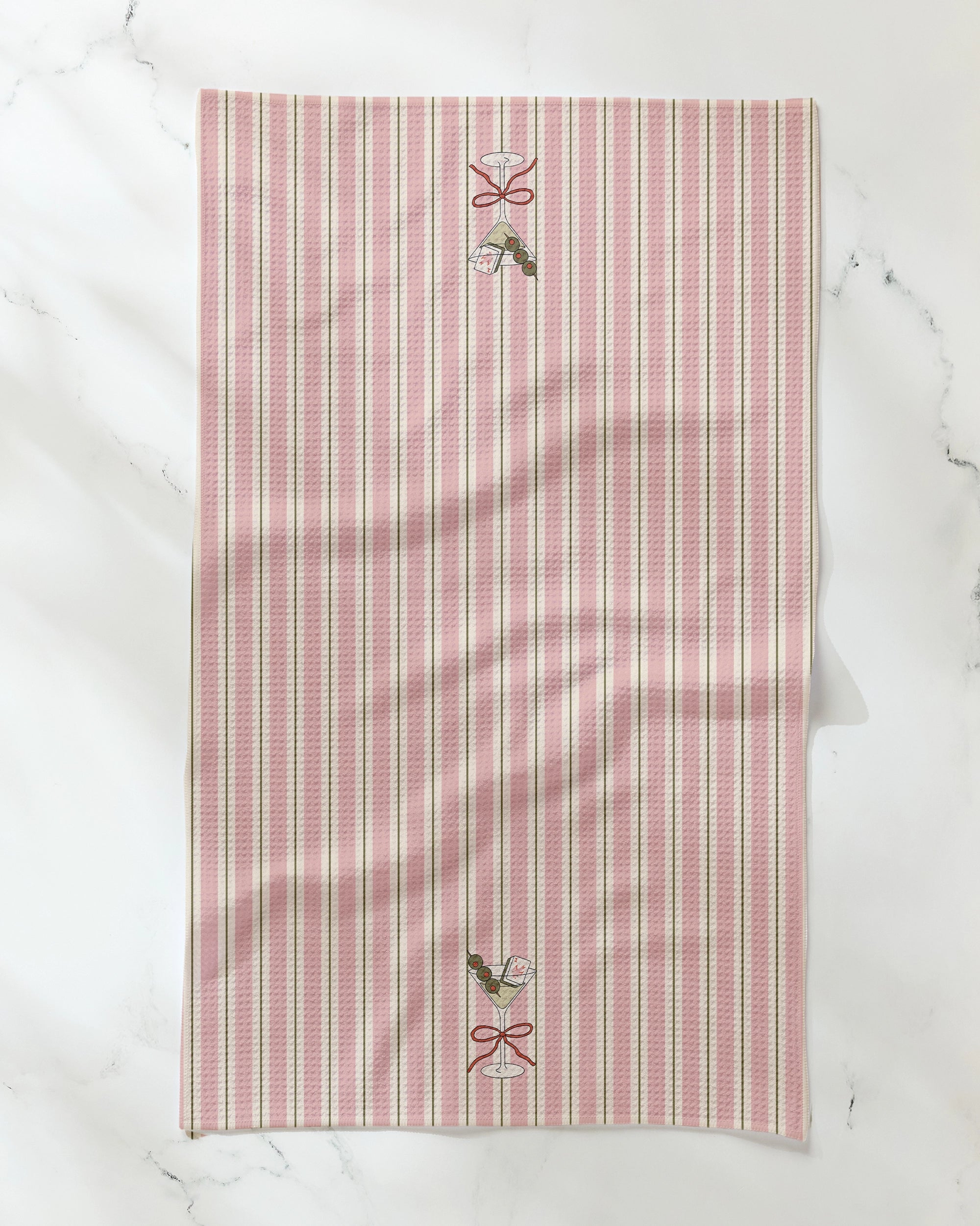 The Geometry Martini Stripe recycled kitchen towel features vertical pink, white, and beige stripes and is adorned with two small holly sprigs tied with red ribbons near the top and bottom edges, displayed on a white marble surface.