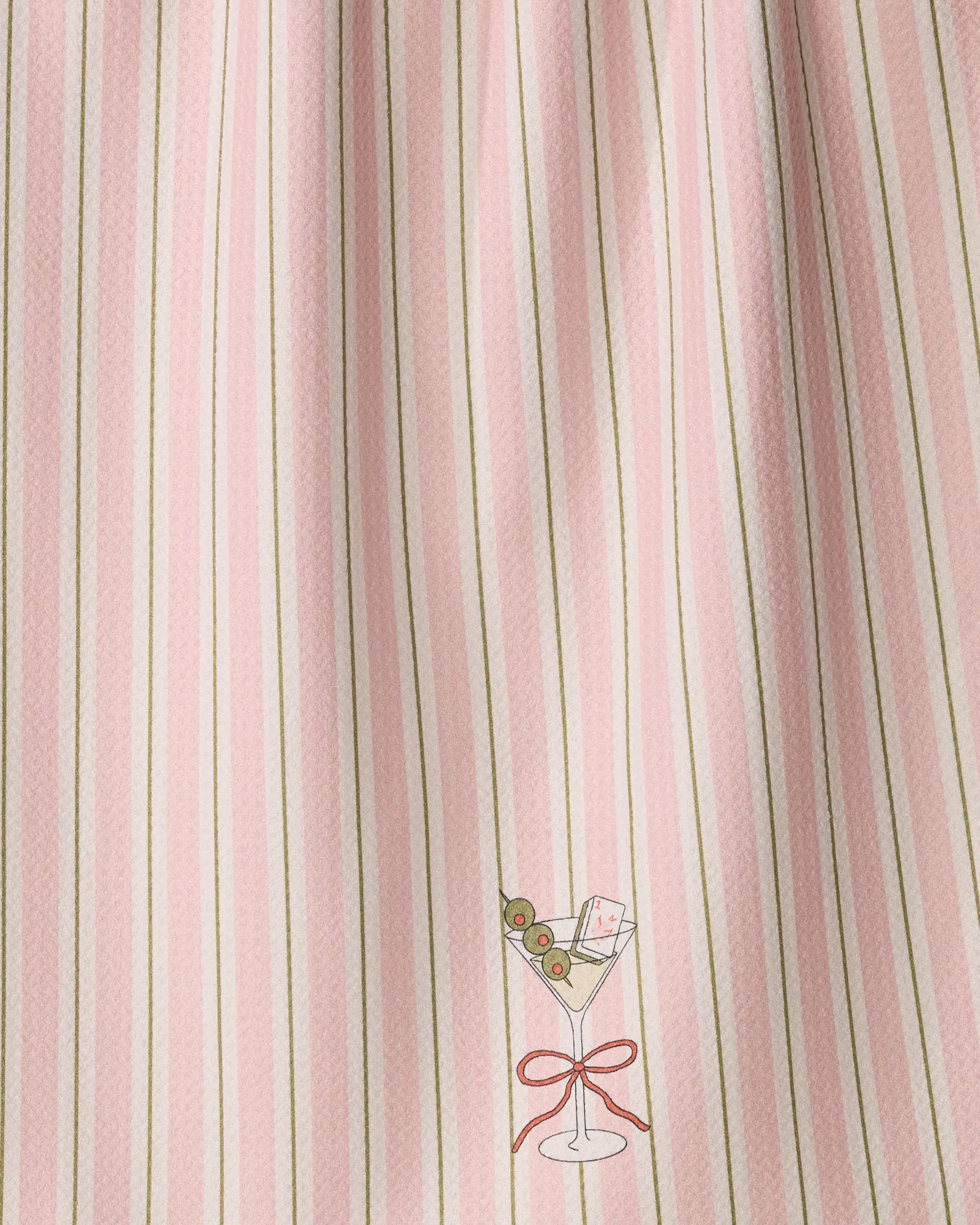 Introducing the Martini Stripe kitchen tea towel by Geometry, featuring a fabric with vertical stripes in shades of pink, white, and thin sage green. Near the bottom, a small illustrated martini glass is adorned with three green olives on a pick and a white rectangular garnish tied with a red ribbon at the base. Crafted from recycled materials, this charming tea towel blends eco-conscious practicality with stylish design.
