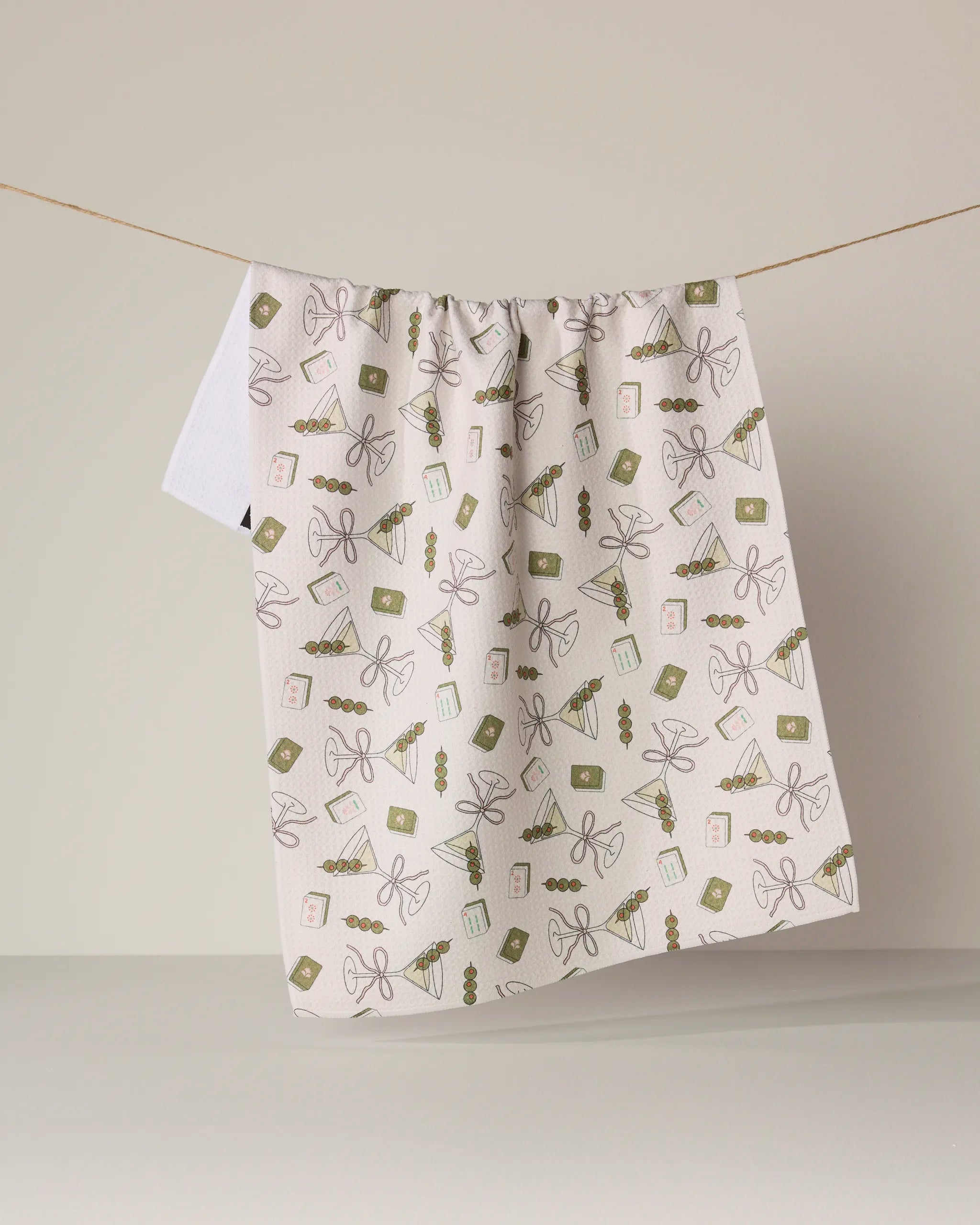 A white kitchen towel from Geometry, called “Martini Tiles,” featuring green and brown gift-themed designs and made from recycled materials, hangs on a string against a plain beige background.