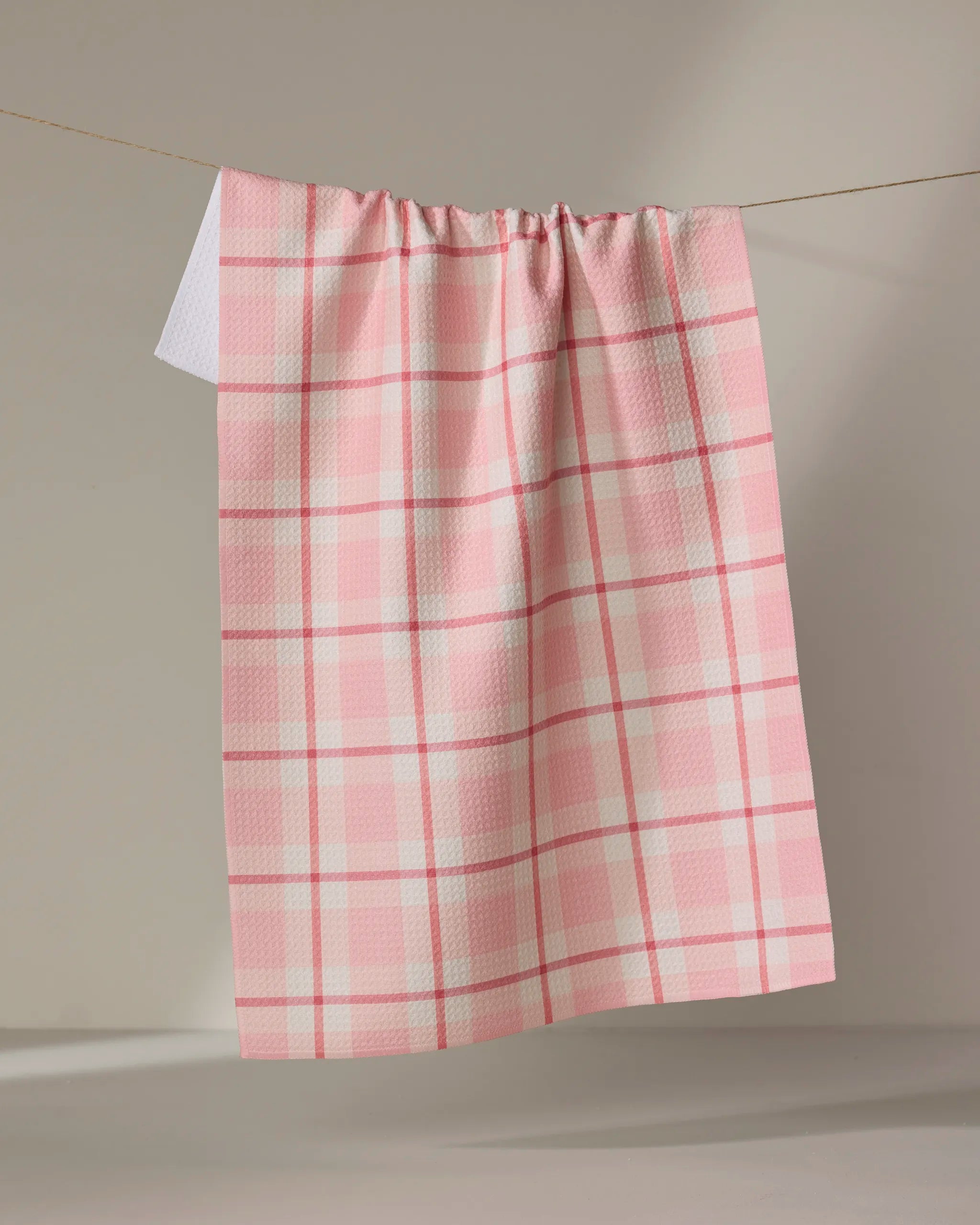 The Marva Lu tea towel by Kelsi Fullmer, featuring a pink and white plaid Geoweave fabric, hangs on a clothesline against a plain light background.