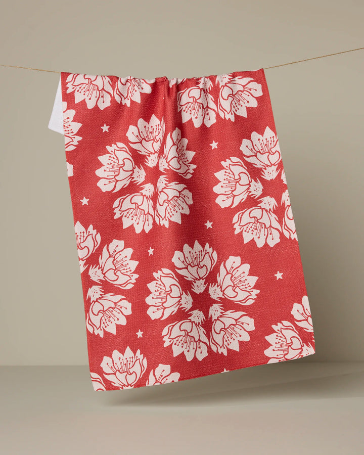 The Maverick Floral tea towel by Geometry, featuring a red background with white floral and star patterns, hangs on a thin string against a beige backdrop.