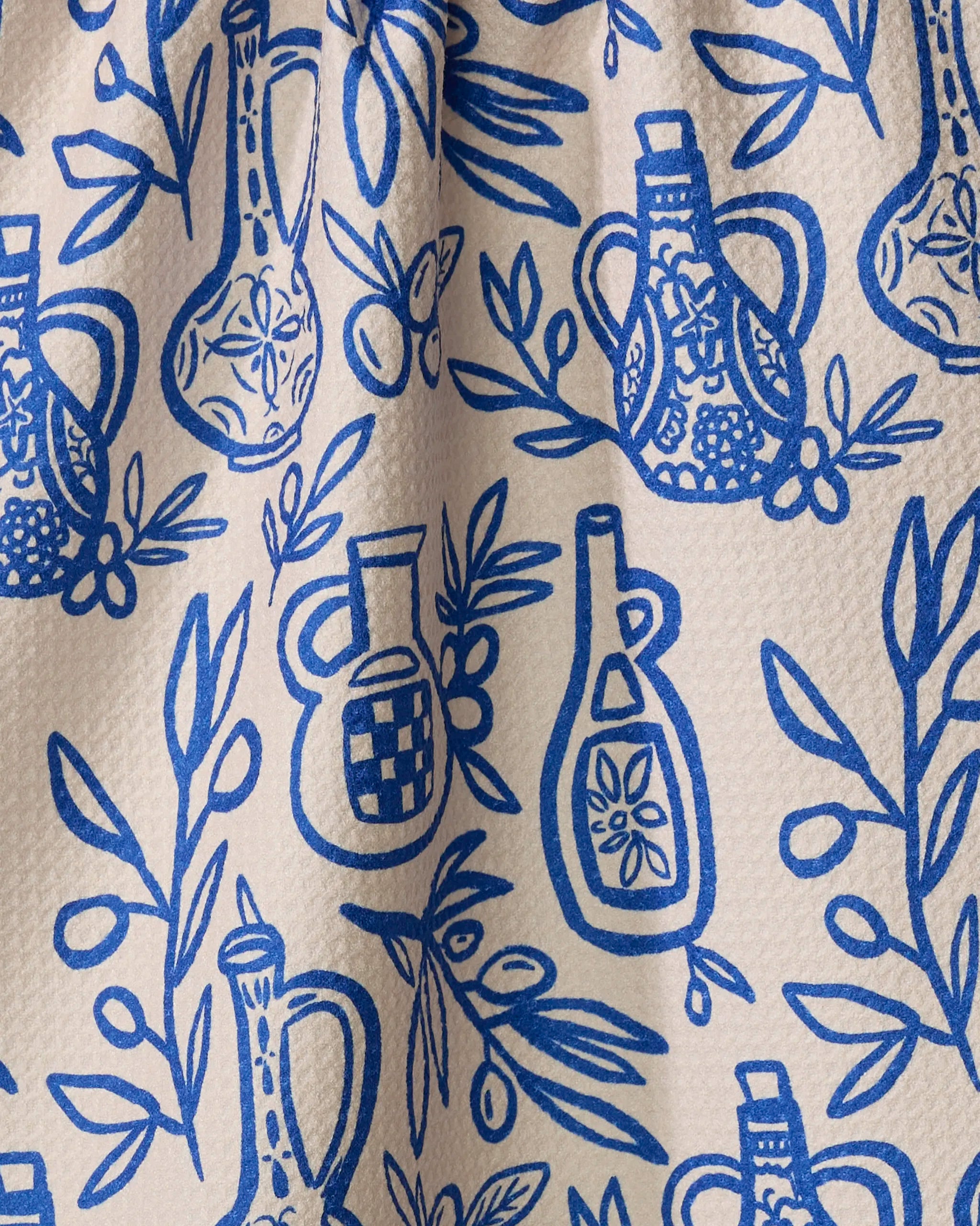 A close-up of Lisa Nelson's "Mediterranean Pour" eco-friendly kitchen towel, featuring a blue and white pattern of olive oil bottles, olive branches, and leaves on a textured cream background.