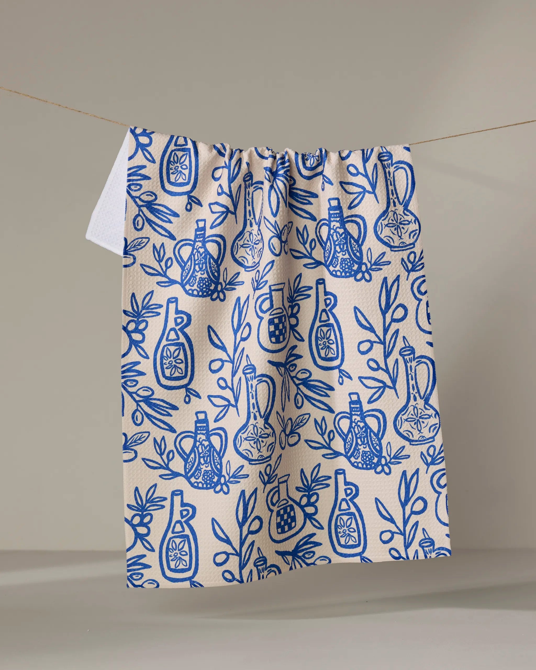The "Mediterranean Pour" kitchen towel by Lisa Nelson, cream-colored and recycled, features blue olive oil bottles, pomegranates, and leafy branches illustrated on it, hanging against a plain beige background.