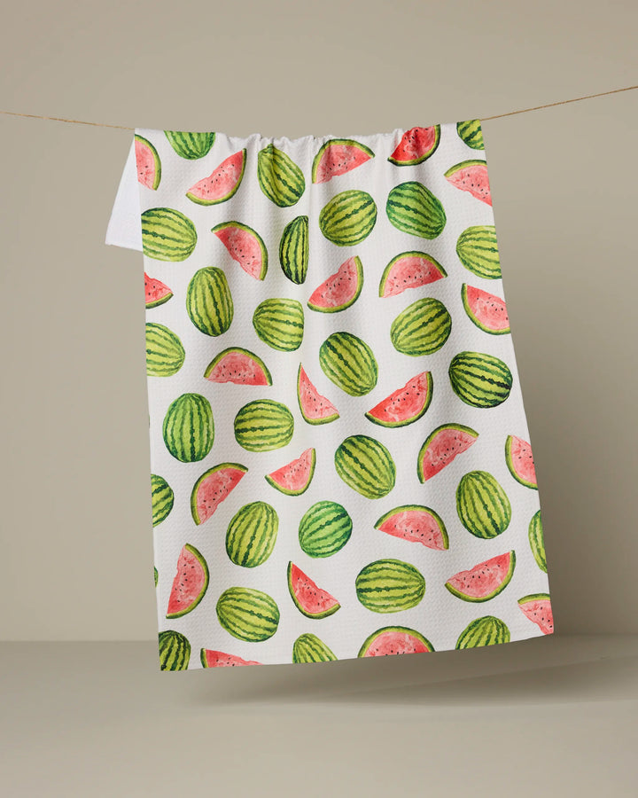 The Melon Market tea towel by Geometry, featuring eco-friendly material and a green and pink watermelon pattern, hangs on a clothesline against a plain beige background.