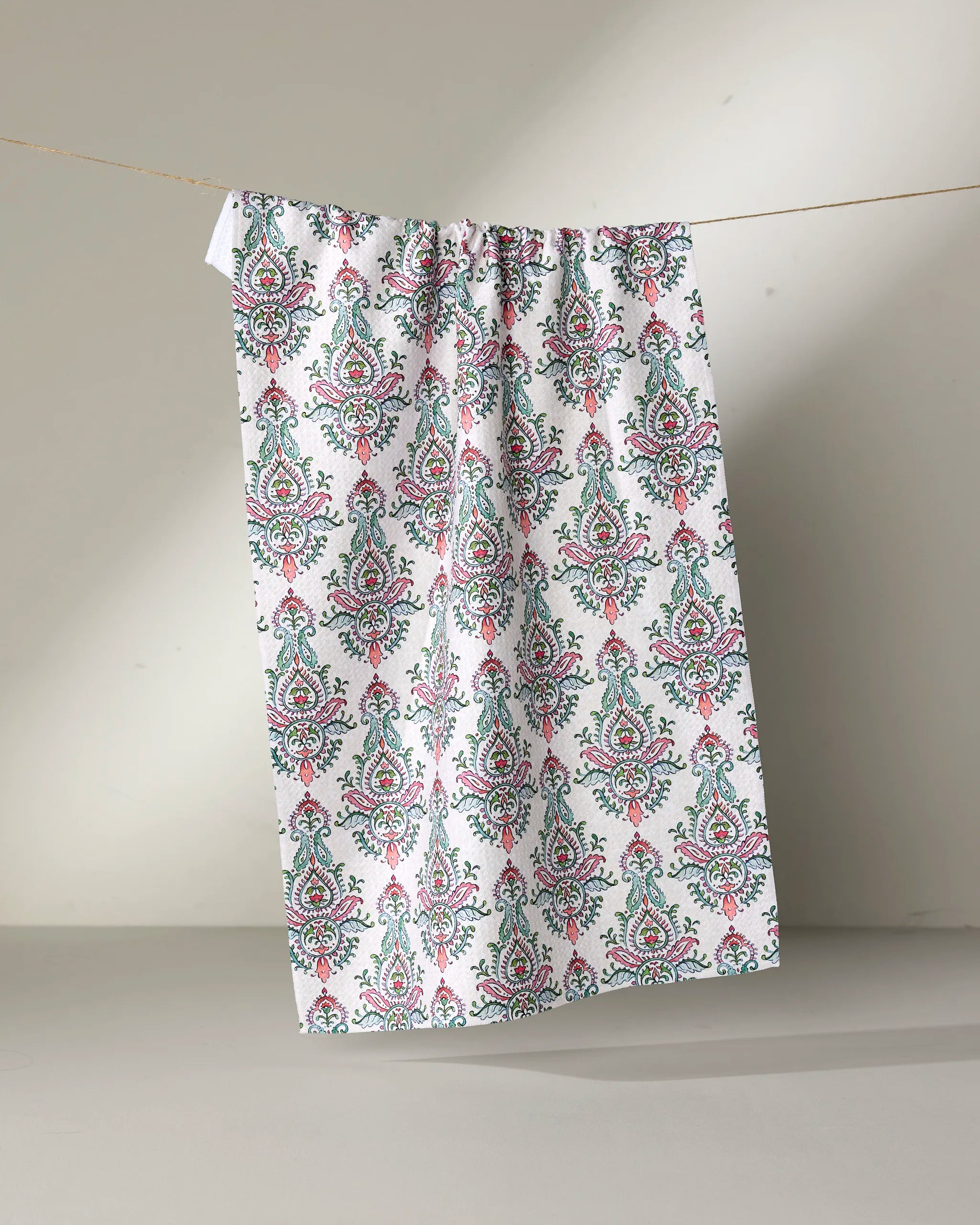 The Memento tea towel by Maayan Alper-Swan, featuring a vibrant green, pink, and blue floral paisley design on white, hangs on a thin line against a plain background.