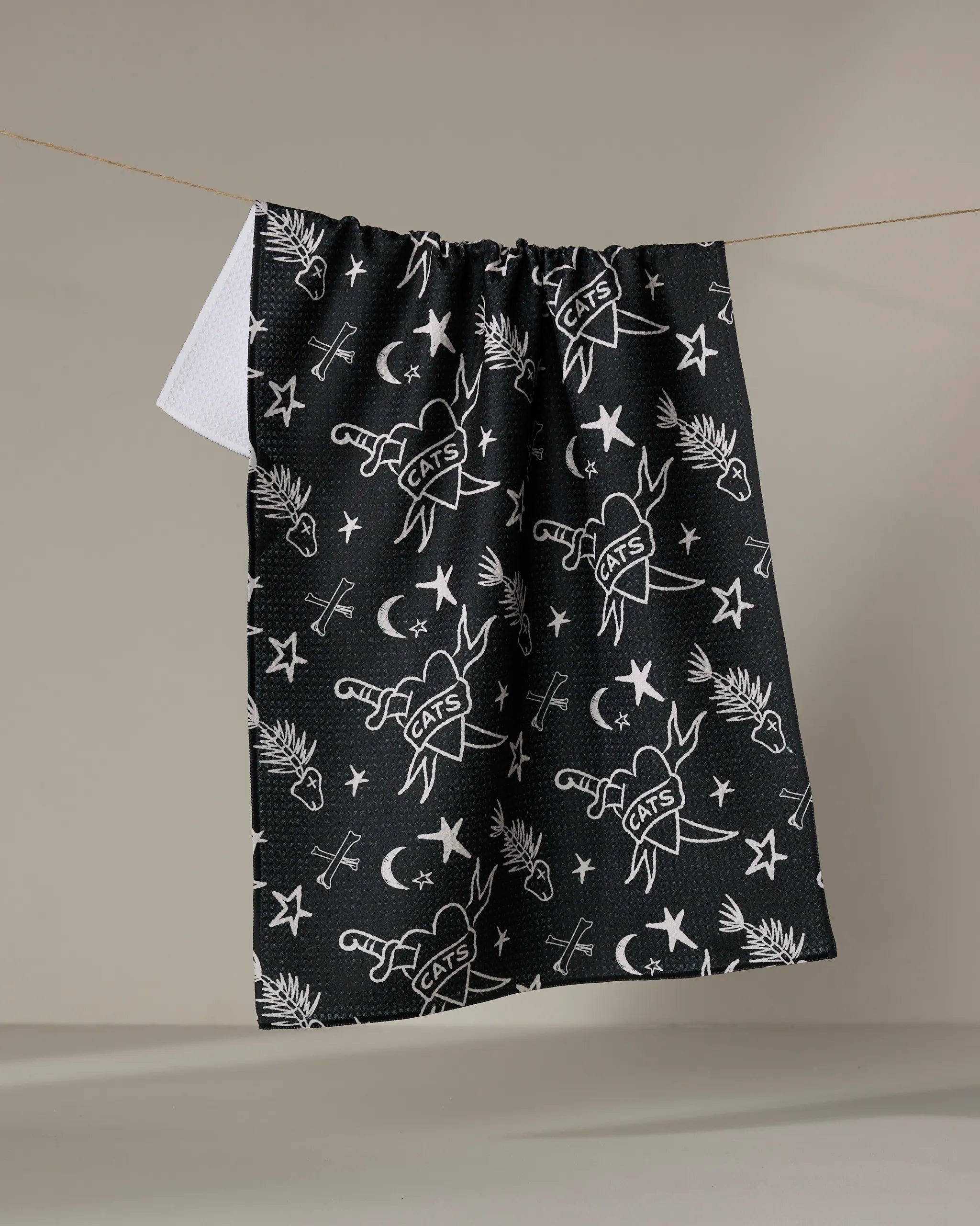 The Cole Strem "Meow or Never" kitchen tea towel, made from recycled materials and featuring black and white stars, moons, eyes, palm trees, and CATS banners, hangs on a line against a neutral background.