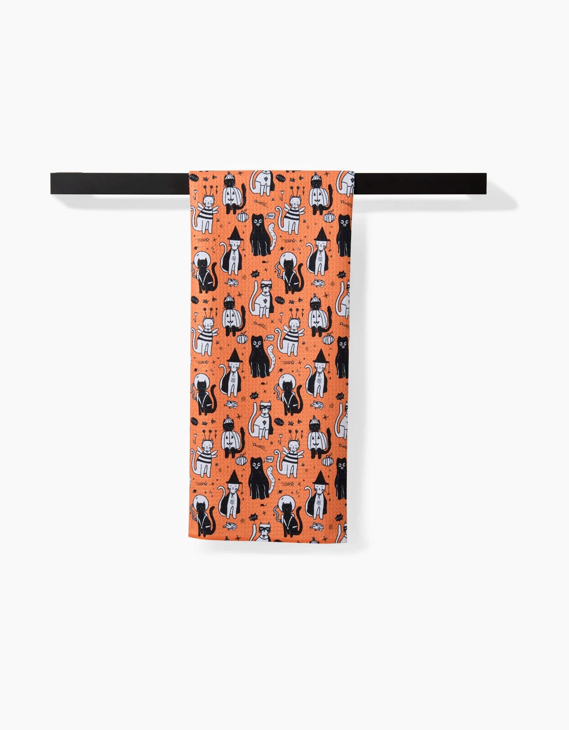 The Meowlloween tea towel by Elisabeth Wing, made from recycled materials, showcases cartoon black and white cats with stars on bright orange. It hangs neatly on a black towel bar against a crisp white background.