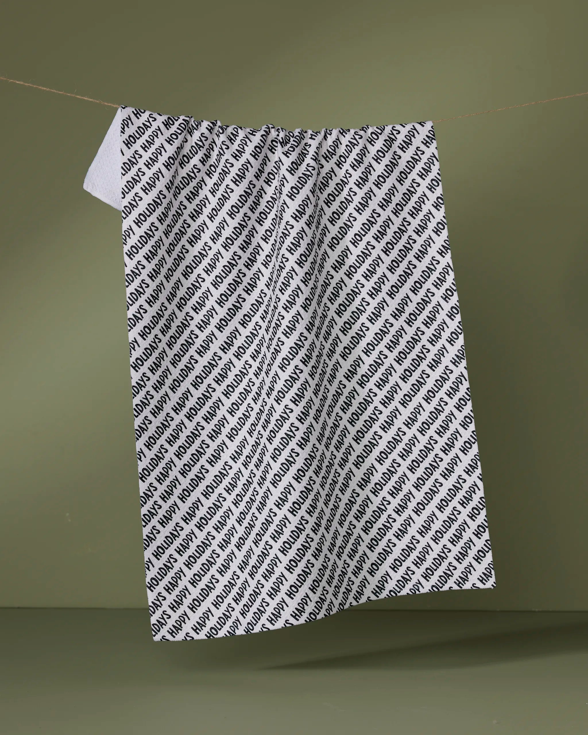 The Merrier tea towel by Geometry is a quick-drying, white towel made from recycled materials, featuring “MONDAY” in black diagonal print, shown hanging on a clothesline against a plain green background.