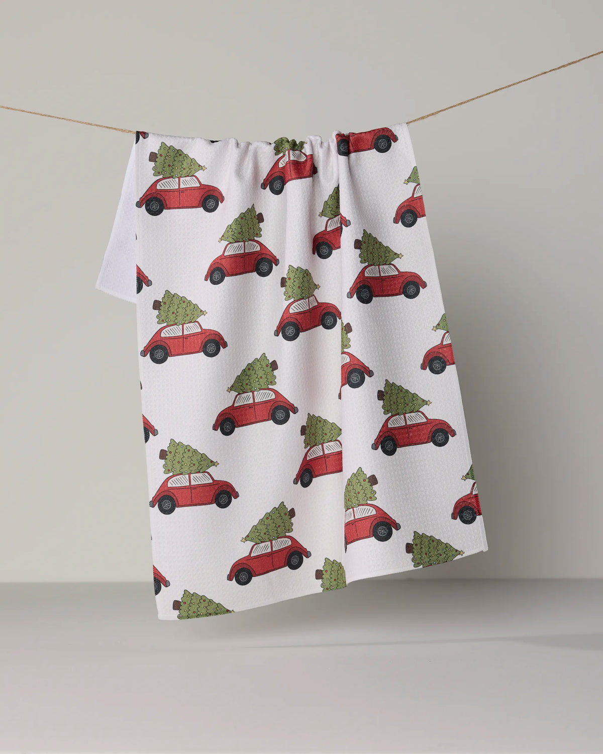 A white kitchen towel from Geometry, called “Merry Miles,” hangs on a clothesline, showcasing a repeating pattern of red cars with green Christmas trees on their roofs. Made from recycled materials, this festive tea towel brings an eco-friendly flair to your holiday décor.