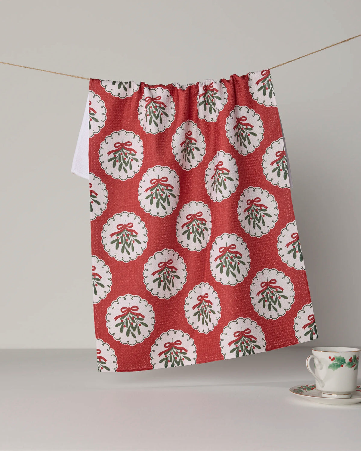 The “Merry Mistletoe” tea towel by Geometry, crafted from recycled materials, features a festive red design adorned with repeating white circles—each showcasing mistletoe tied with a red bow. Ultra-absorbent and charming, it hangs on a string to brighten your kitchen. Beneath it, a floral teacup and saucer rest on a white surface.