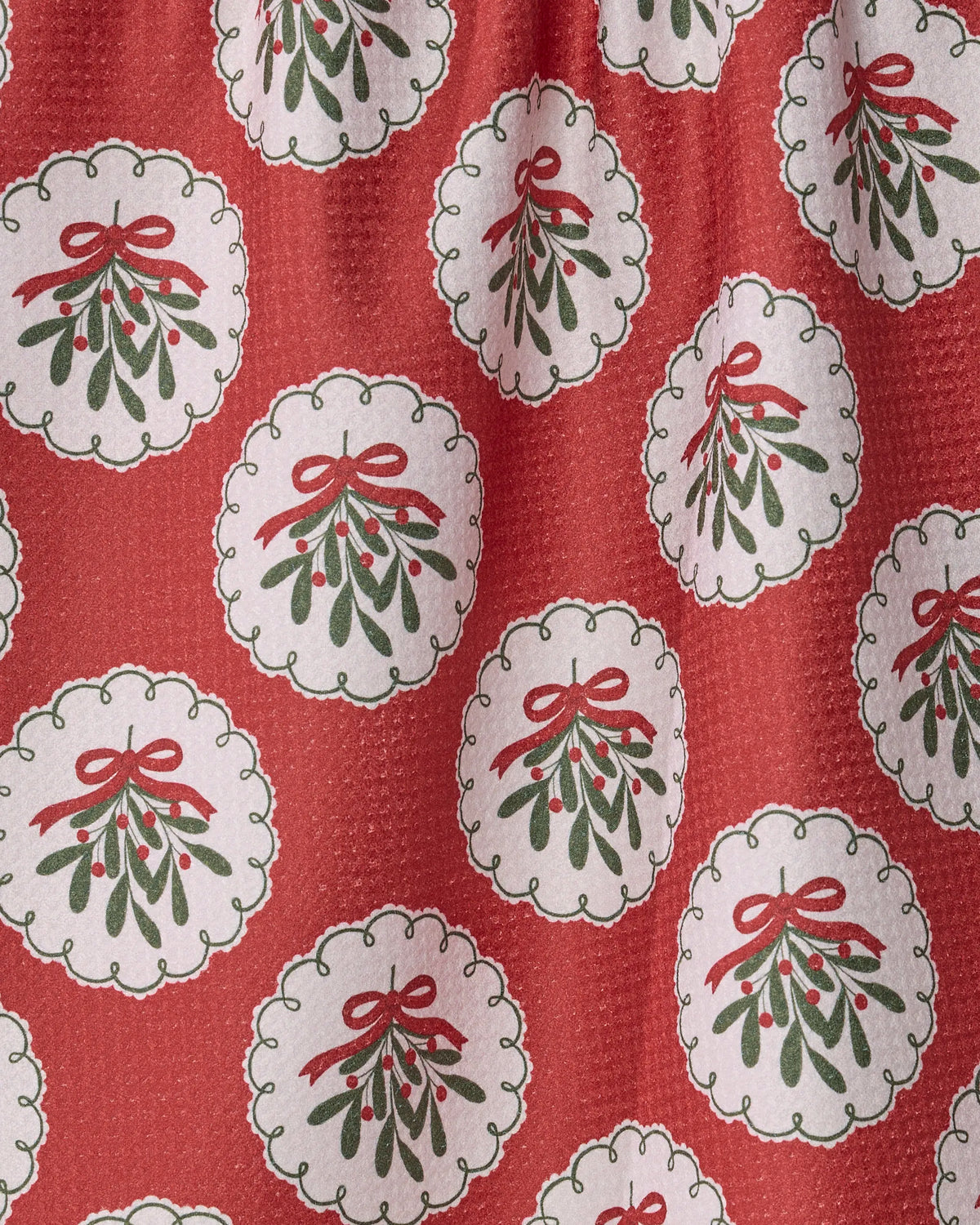 The Geometry Merry Mistletoe kitchen towel features a red fabric adorned with a repeating pattern of white scalloped circles, each showcasing green mistletoe accented by red berries and a bright red bow. The textured background adds depth and character, making this ultra-absorbent towel both festive and functional. Crafted from recycled materials, it’s a cheerful addition to your home for the holidays.