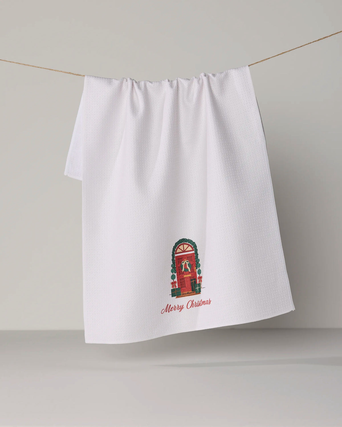 Bring festive cheer to your kitchen with the Geometry ‘Merry Welcome’ tea towel. This white, ultra-absorbent towel is crafted from recycled materials and features an illustration of a red door adorned with a holiday wreath and garland, set against a simple gray background. Below the door, “Merry Christmas” appears in red for that perfect seasonal touch.