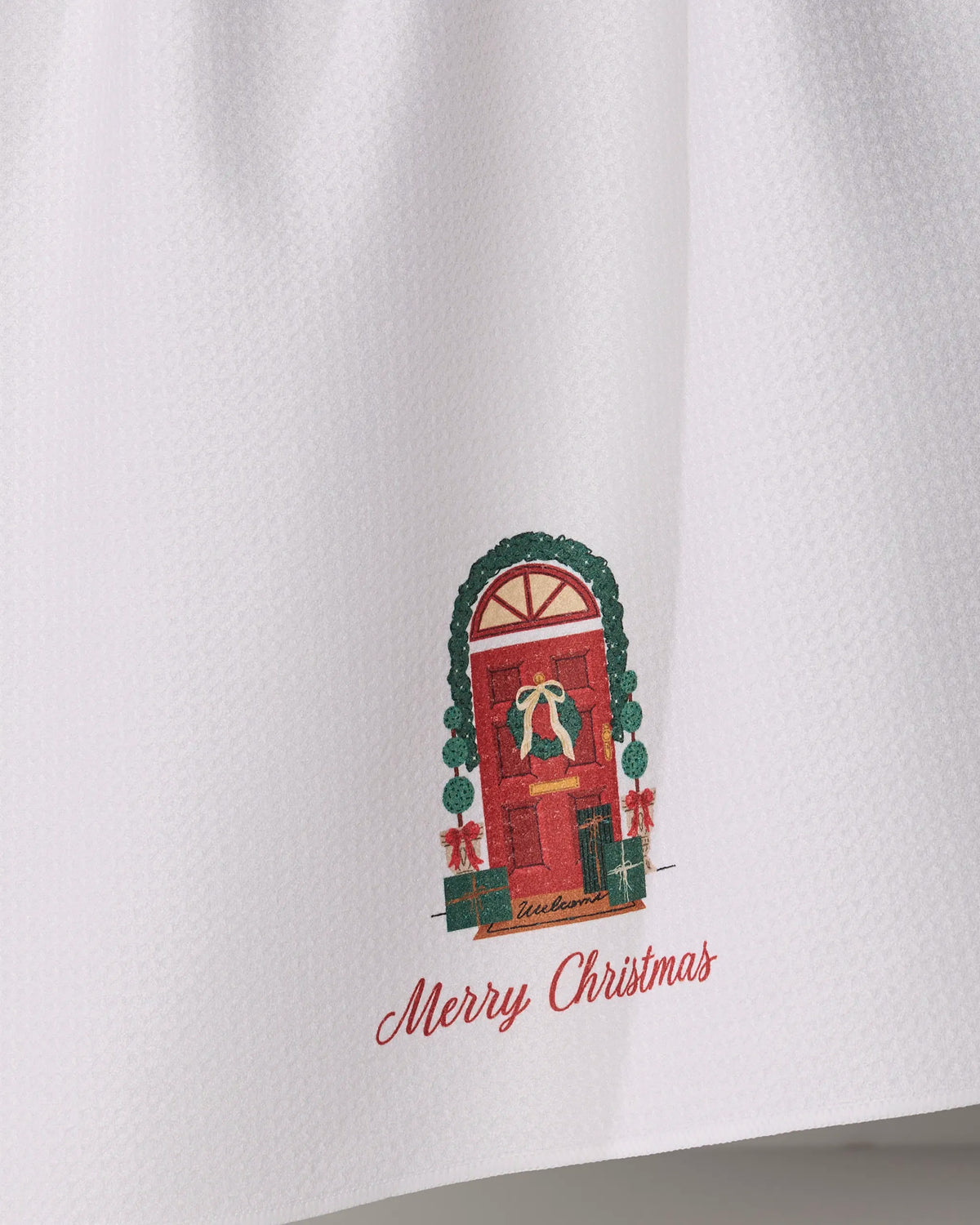 Introducing the “Merry Welcome” tea towel by Geometry—a white, ultra-absorbent kitchen essential adorned with an illustration of a red door beautifully decorated with a wreath, garland, and festive gifts. The cheerful phrase “Merry Christmas” is elegantly scripted in red below the scene, making this charming towel both functional and perfect for adding holiday spirit to your home.