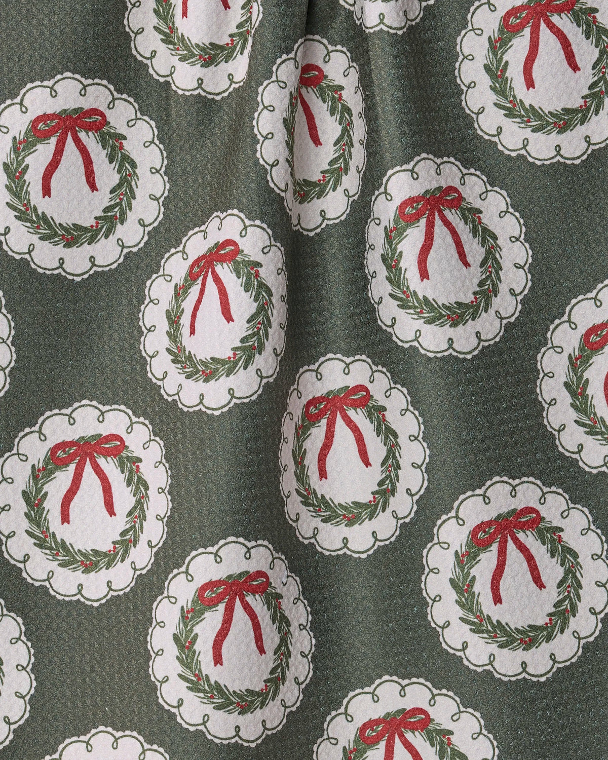 The “Merry Wreaths” tea towel by Geometry features green fabric adorned with a repeating pattern of white circles, each highlighting a Christmas wreath with a red bow. Crafted from recycled materials, this eco-friendly towel offers a textured look with slight folds and ensures quick-drying convenience.