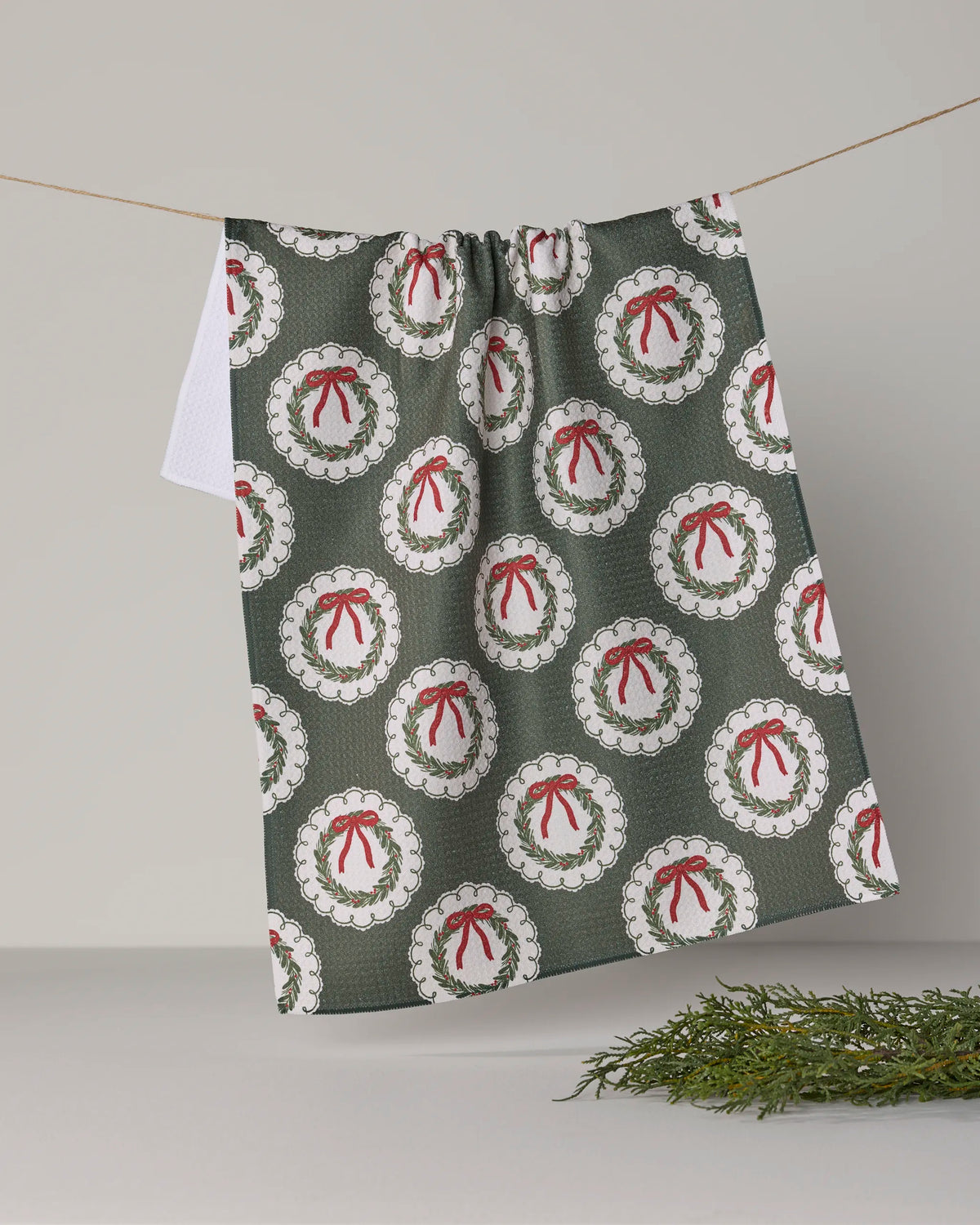 A festive, quick-drying Geometry tea towel called “Merry Wreaths,” featuring a pattern of white holiday wreaths tied with red bows, hangs on a string with a sprig of greenery resting on the light gray surface below.
